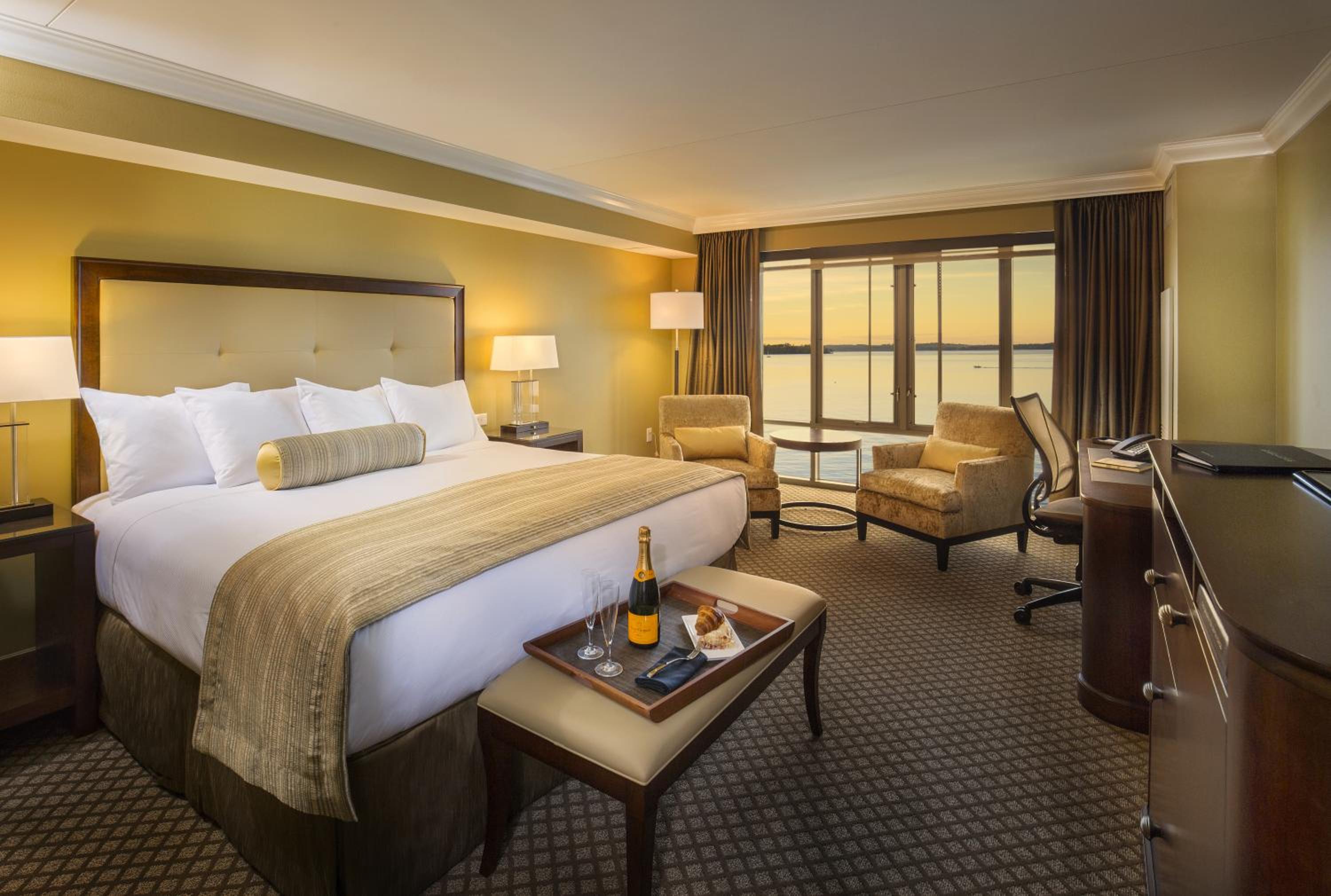 Hotel bedroom with a made bed, champagne tray on an ottoman bench, and armchairs facing lake-view windows.