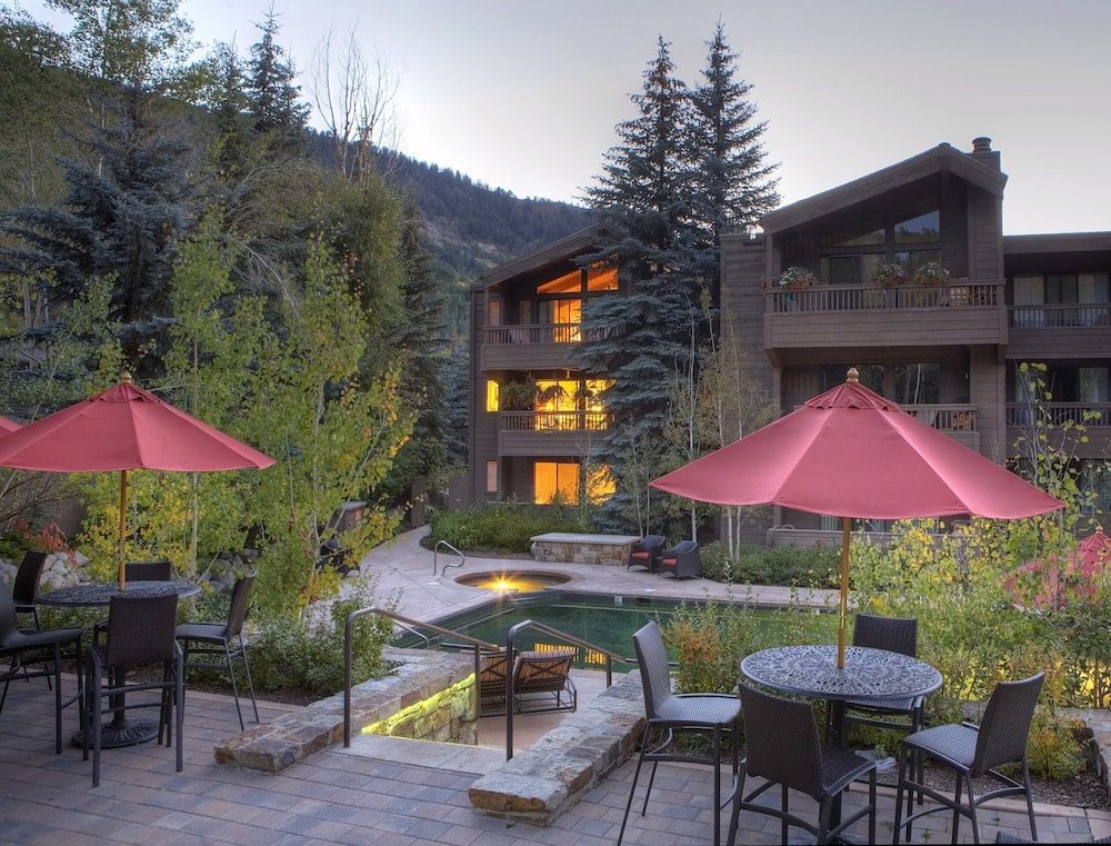Paved terrace with umbrella-shaded patio tables overlooking a swimming pool and lodge exterior at dusk.