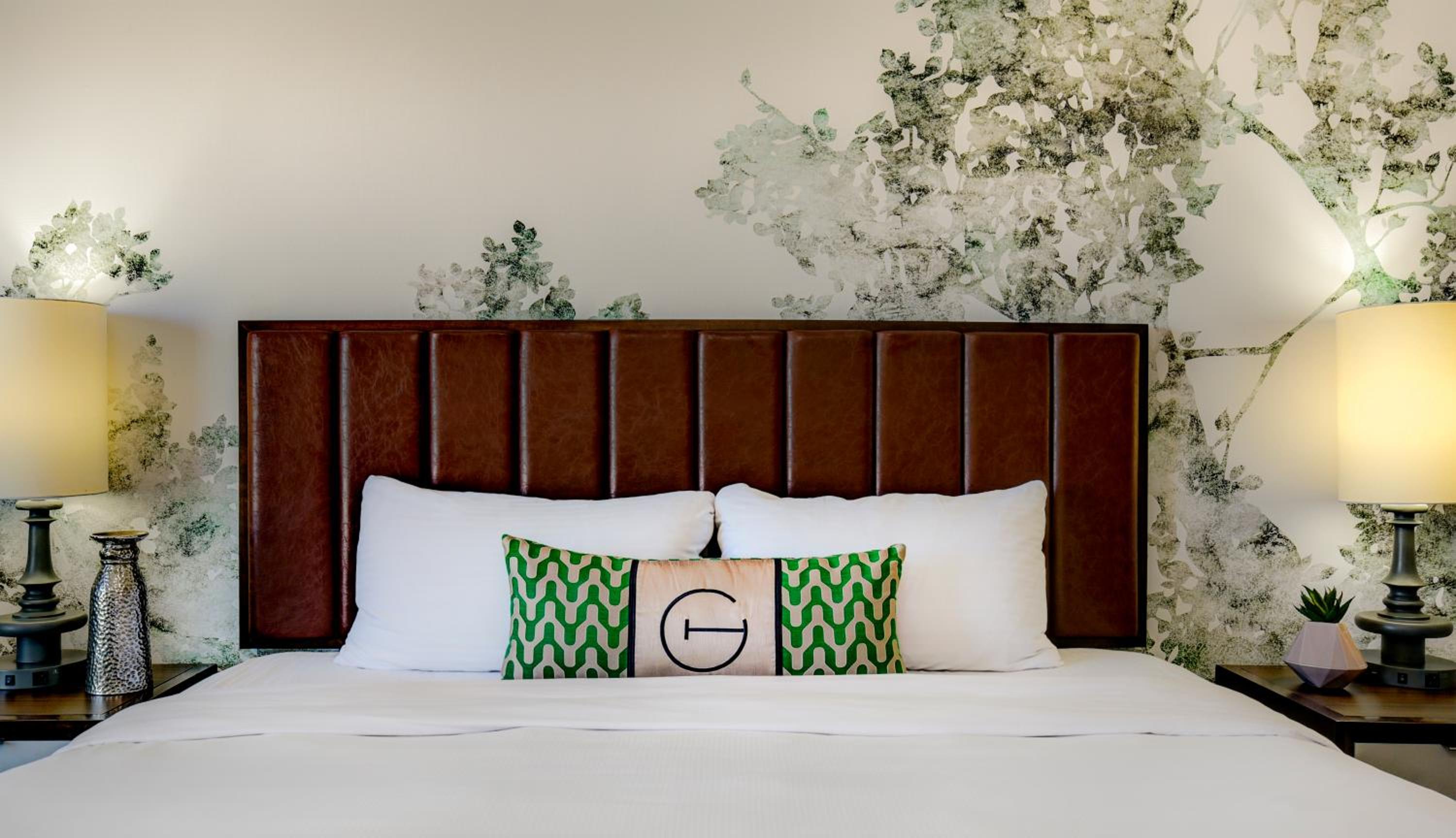 A hotel bed with a brown leather headboard, patterned accent pillow, bedside lamps, and tree-print wallpaper.