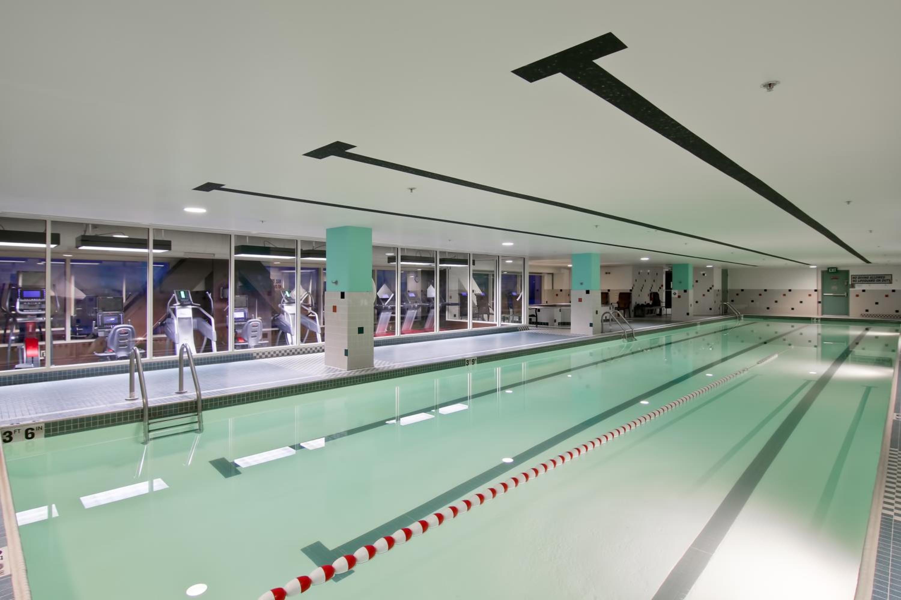 Indoor lap swimming pool with lane markings alongside glass windows looking into a fitness center.
