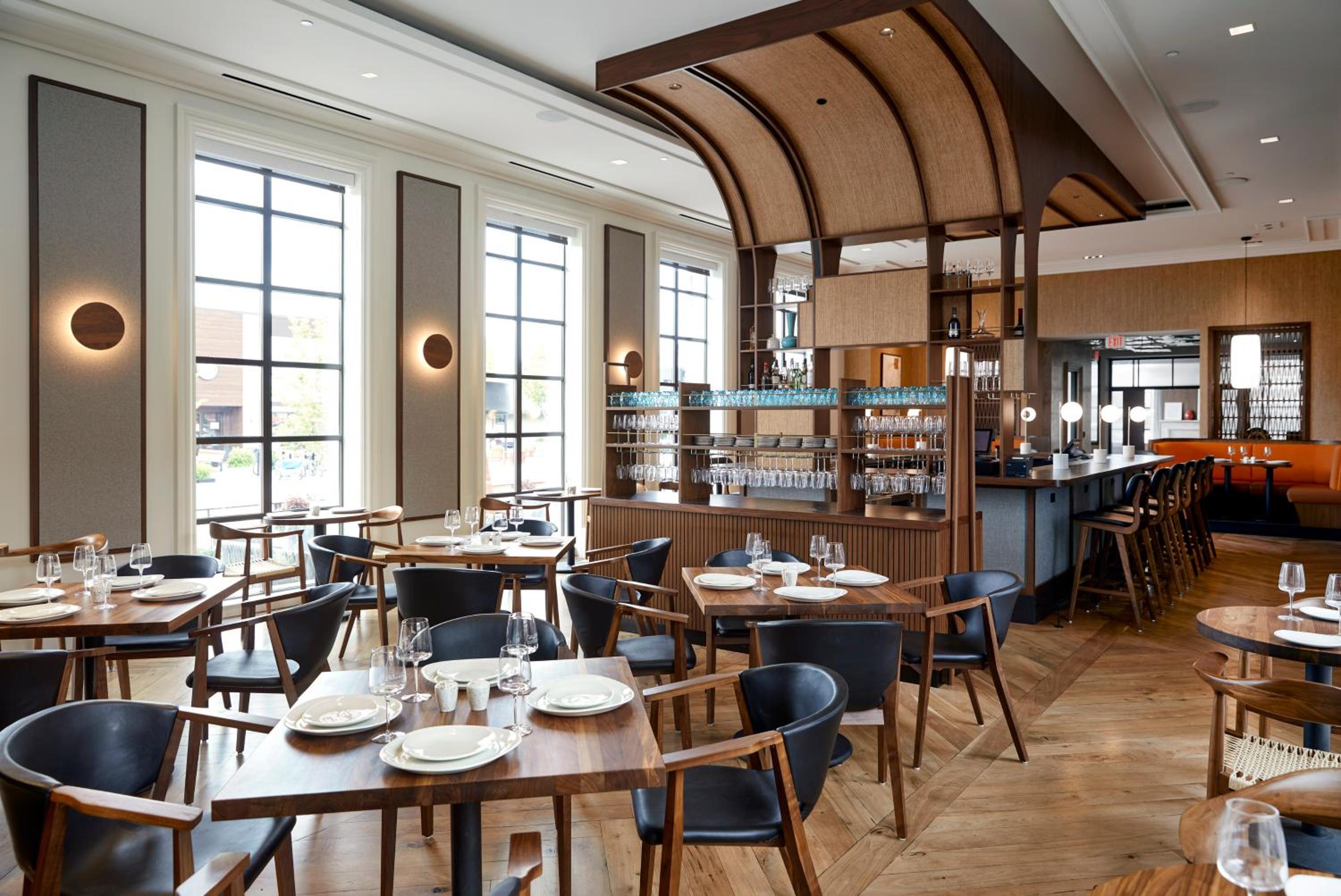 Restaurant dining area with wooden tables, dark leather chairs, tall grid windows, and an arched timber canopy.