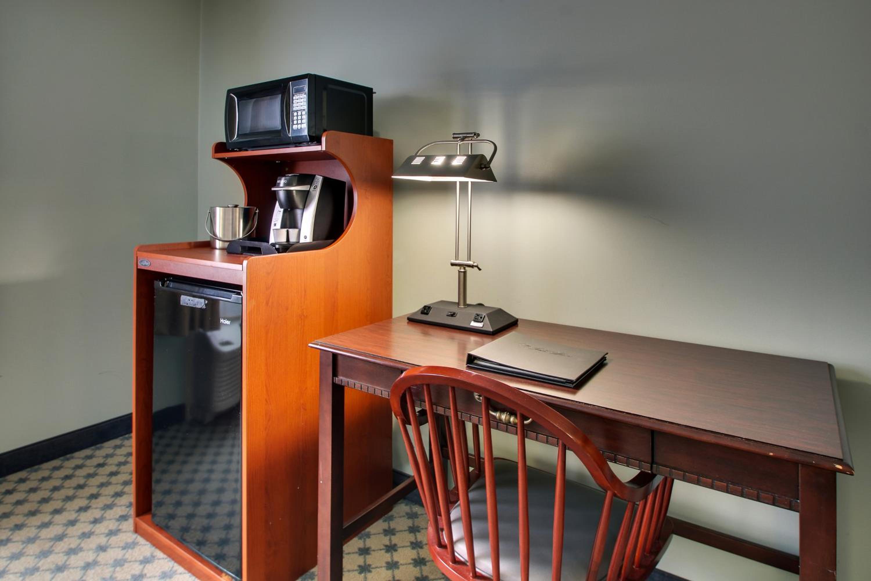A wooden desk with a lamp and chair sits beside a cabinet holding a microwave, coffee maker, and mini fridge.