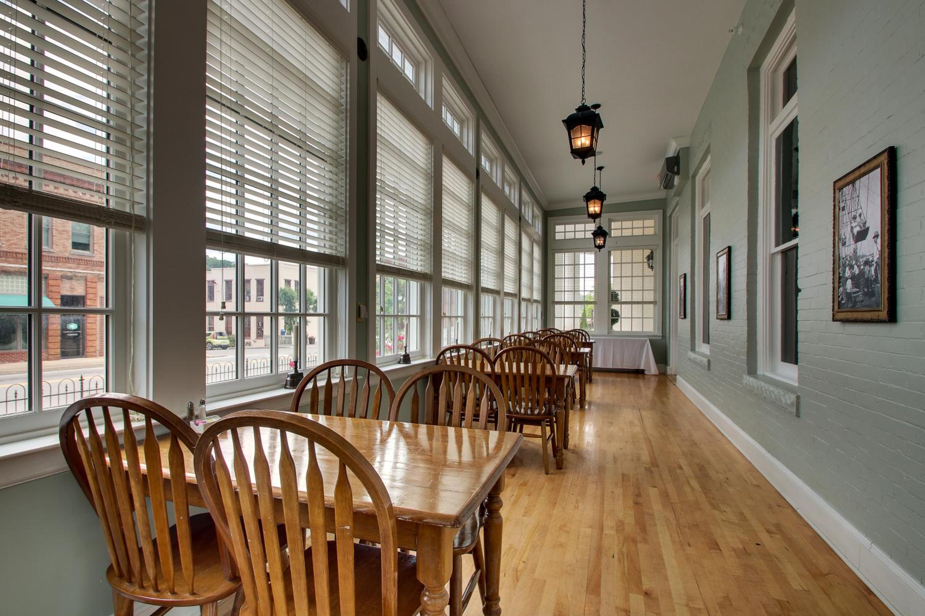 An enclosed sunroom with wooden dining tables, hardwood flooring, and tall windows with blinds.