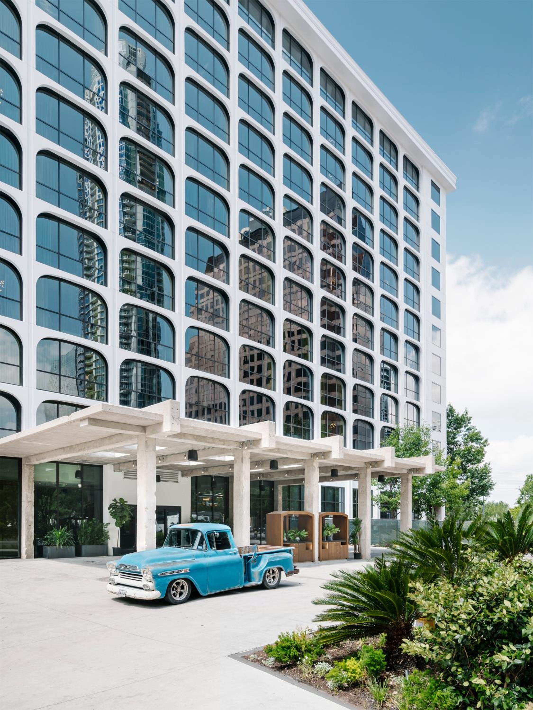 A vintage turquoise pickup truck is parked outside the hotel facade featuring curved white window grids.