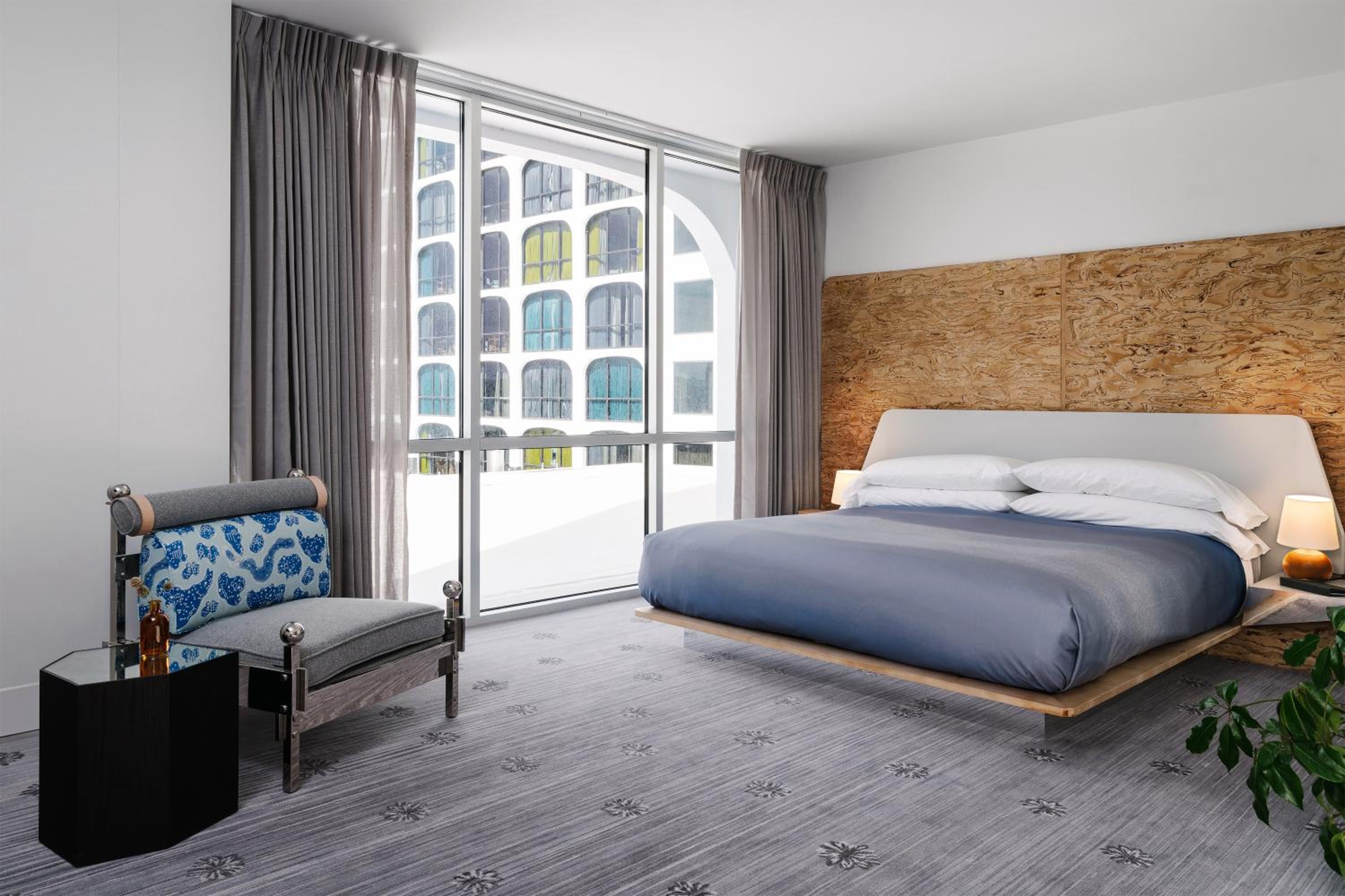 A hotel room contains a low platform bed with a burl wood headboard, an armchair, and large windows.