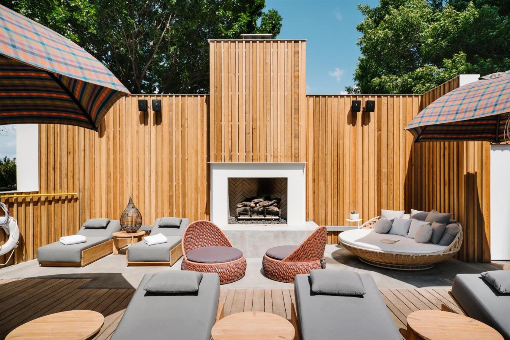 An outdoor terrace features slatted wood walls, a modern fireplace, lounge seating, and plaid umbrellas.