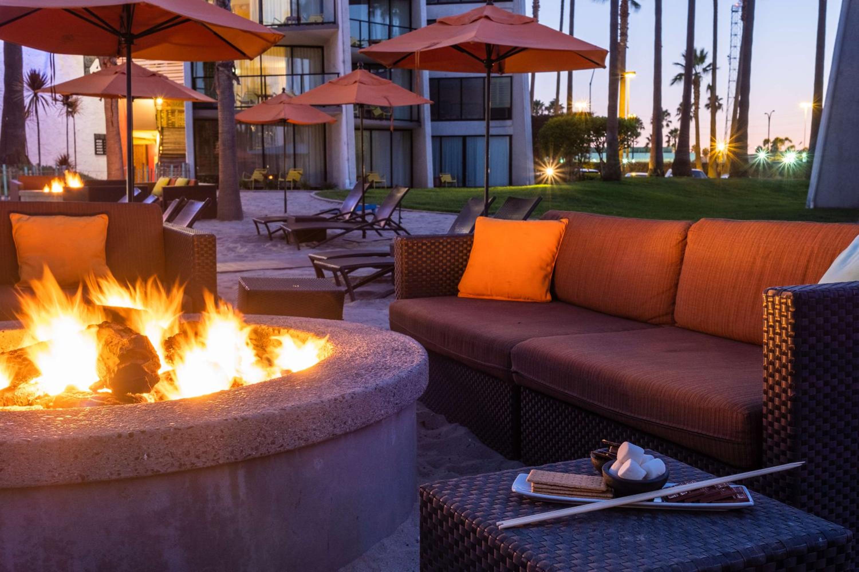 A flaming stone fire pit surrounded by wicker patio seating with orange cushions and umbrellas at twilight.