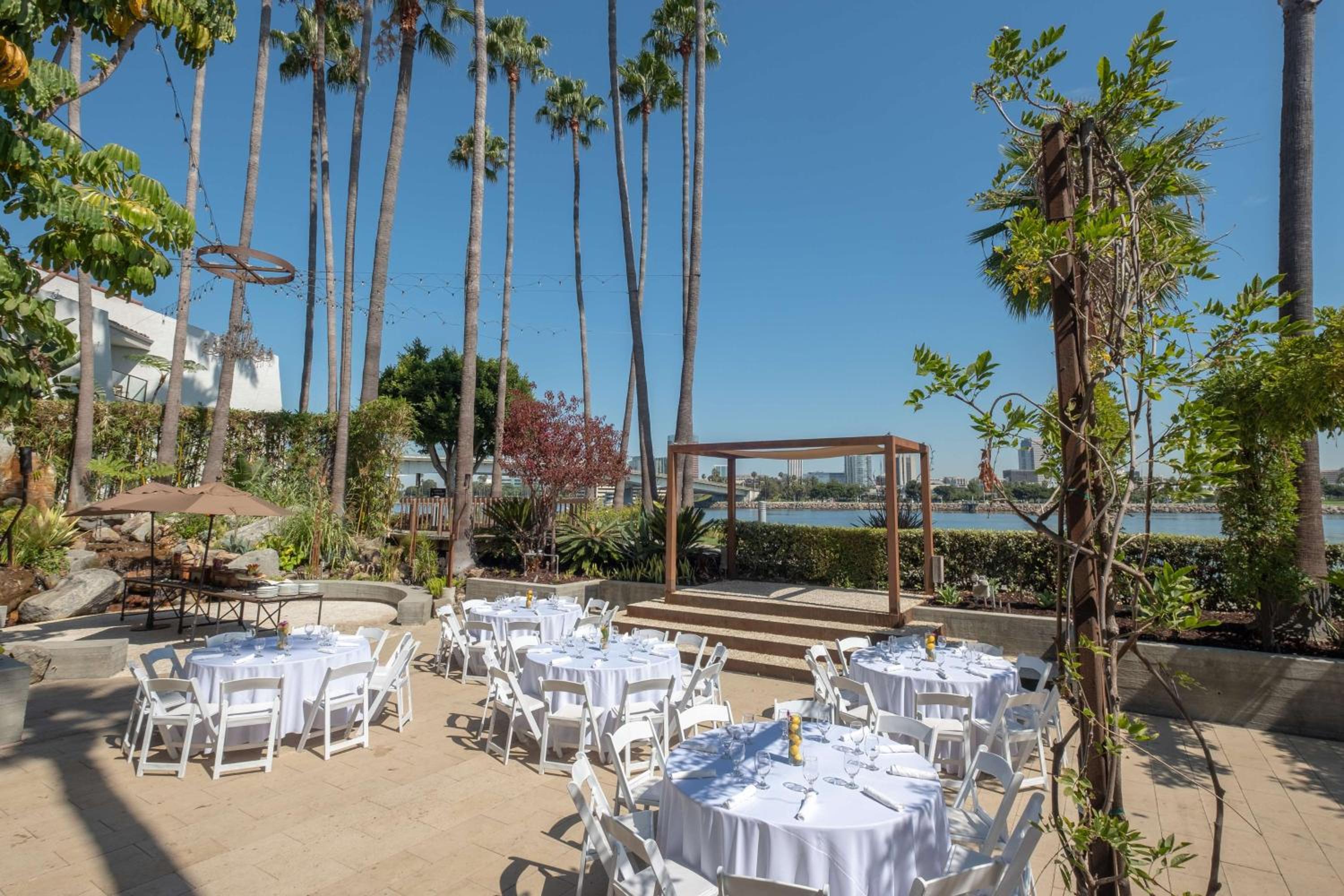 An outdoor paved terrace set with white-clothed tables, a wooden platform, and tall palm trees overlooking the bay.