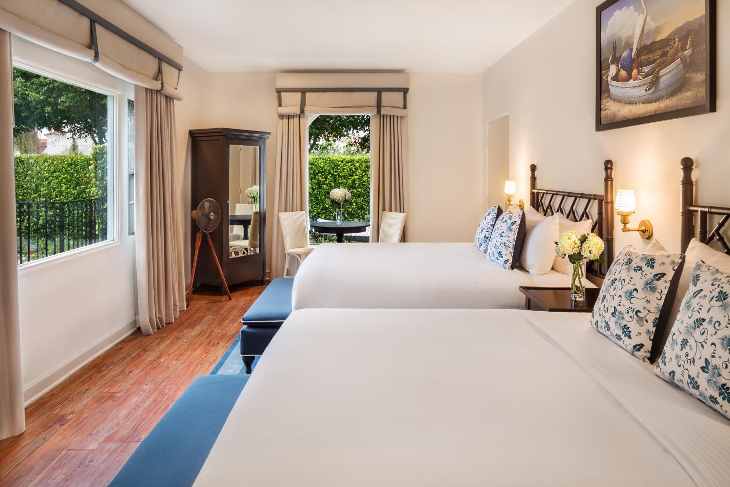 A guest bedroom with beds dressed in white linens, floral accent cushions, wood flooring, and sunlit windows.