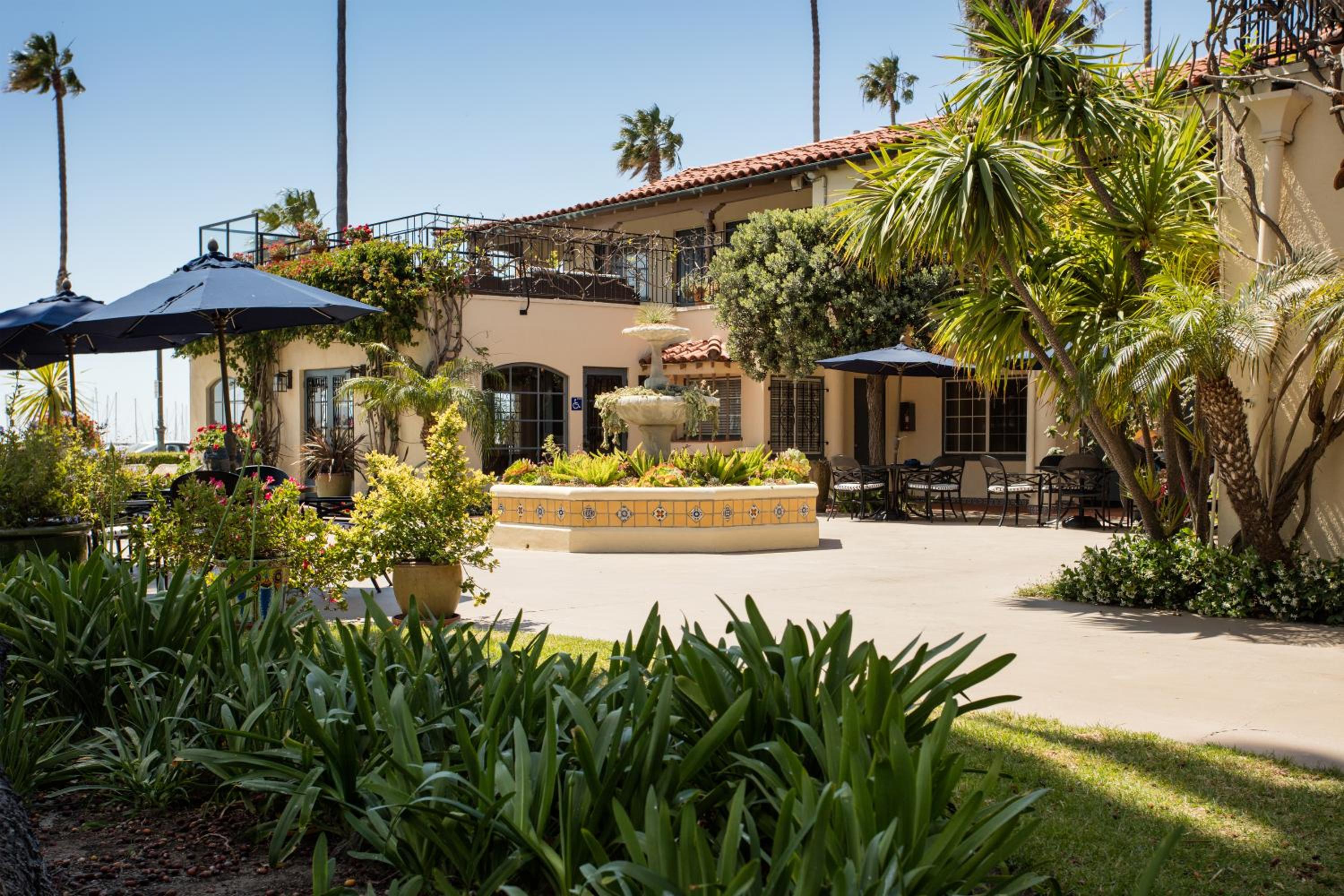 A courtyard patio with a tiled central fountain, umbrella-shaded tables, palm trees, and Spanish-style architecture.