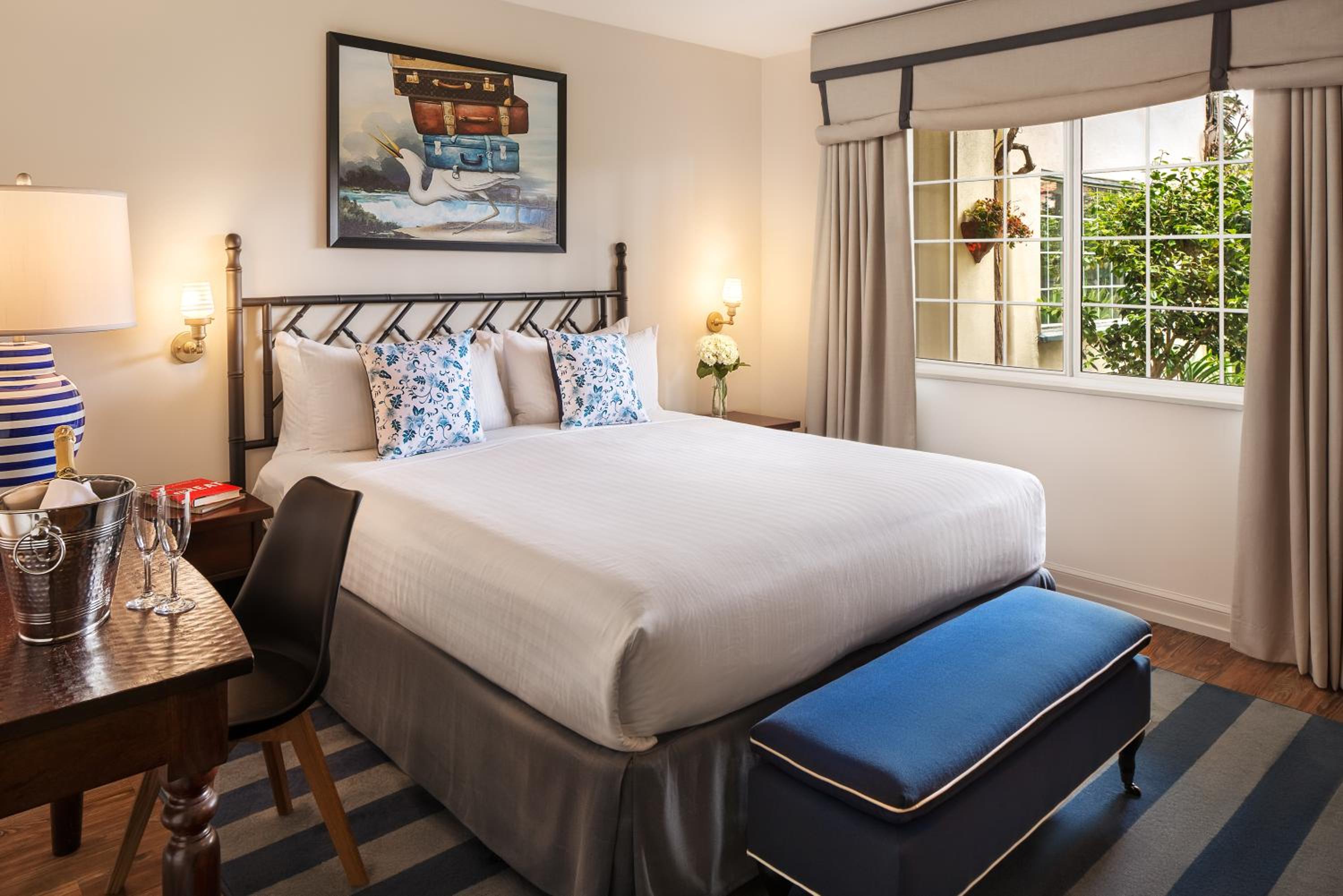 A bedroom featuring an iron headboard, white bedding, an upholstered blue bench, and a window to courtyard greenery.