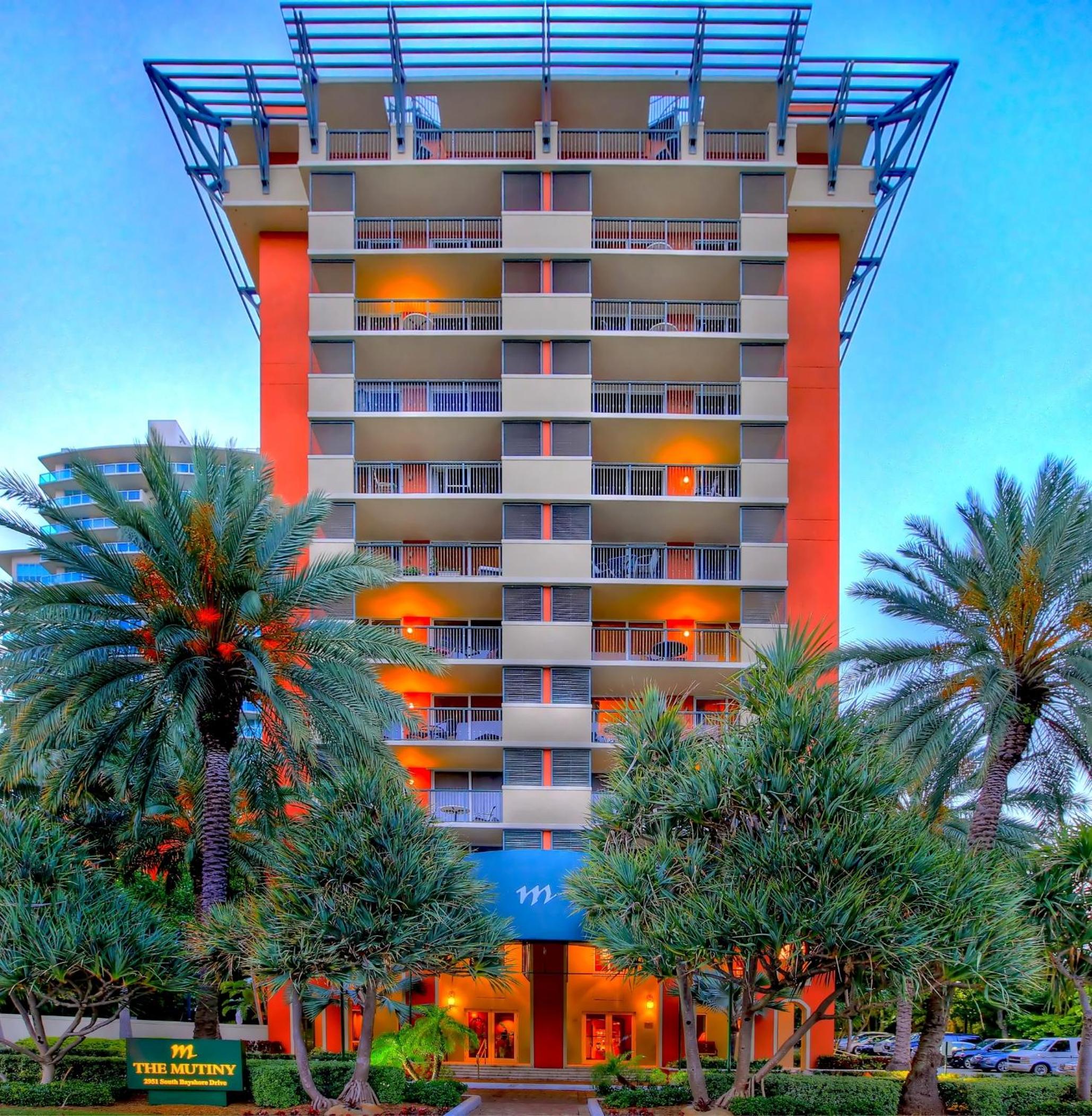 Facade of The Mutiny Hotel with orange accents, balconies, and palm trees flanking the entrance.