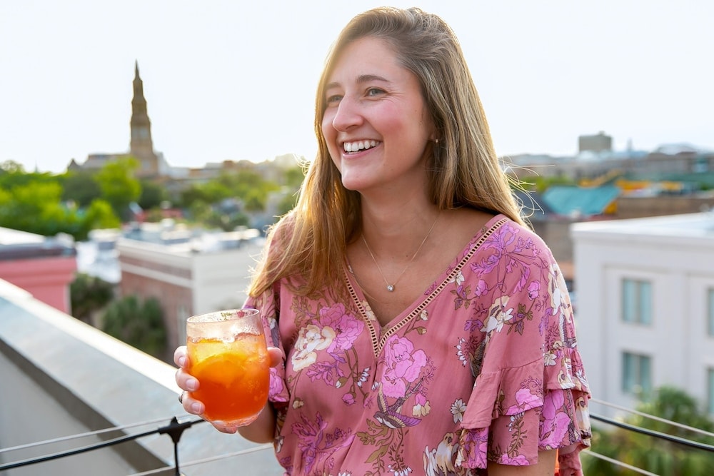 A woman smiles holding a cocktail on a rooftop terrace overlooking the city skyline under daylight.