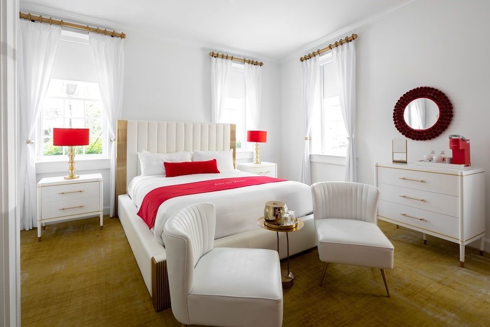A hotel bedroom with a white upholstered bed, red accent pillows, armchairs, and an ochre rug.