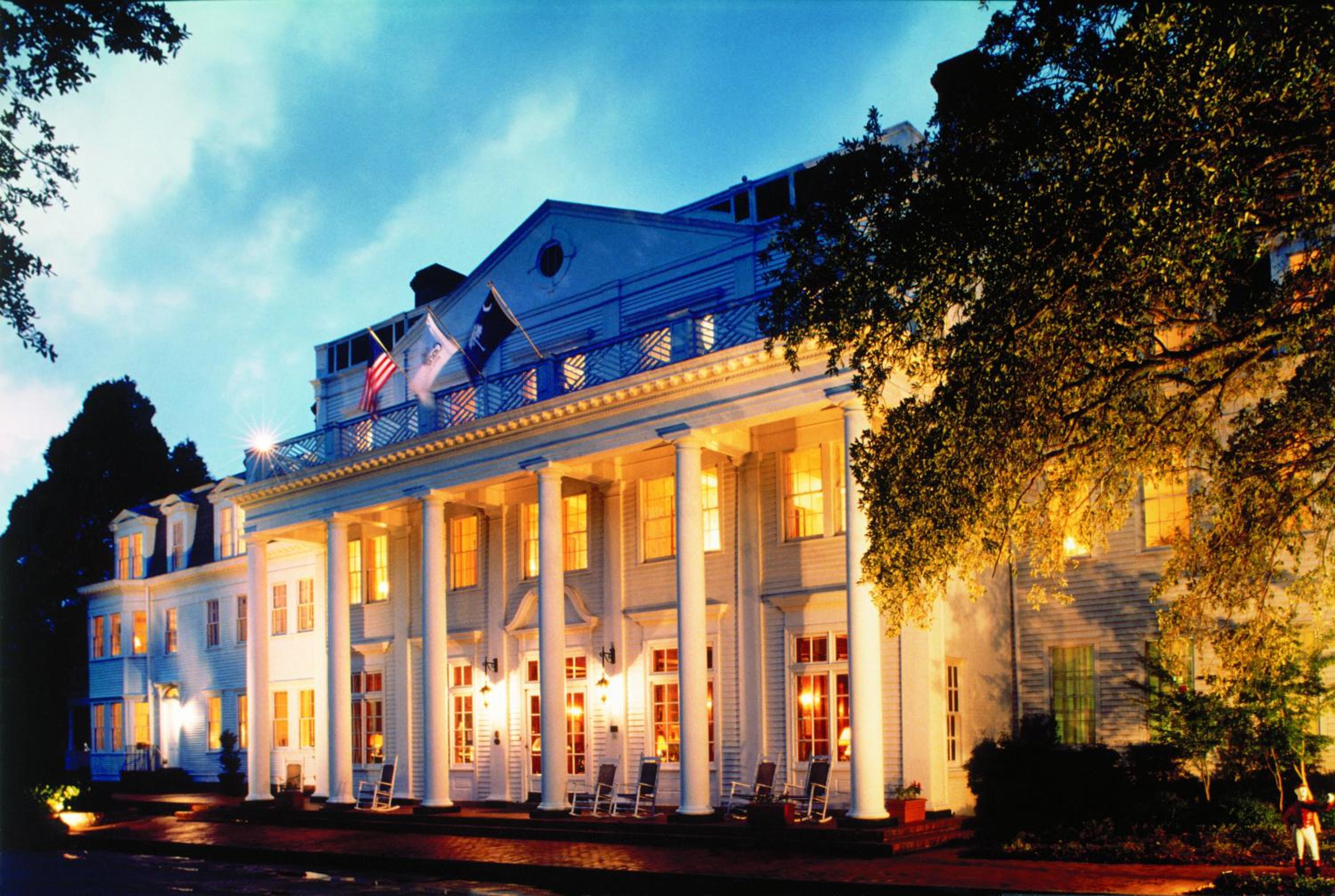 Illuminated white facade of The Willcox at dusk, featuring tall columns, porch rocking chairs, and lit windows.