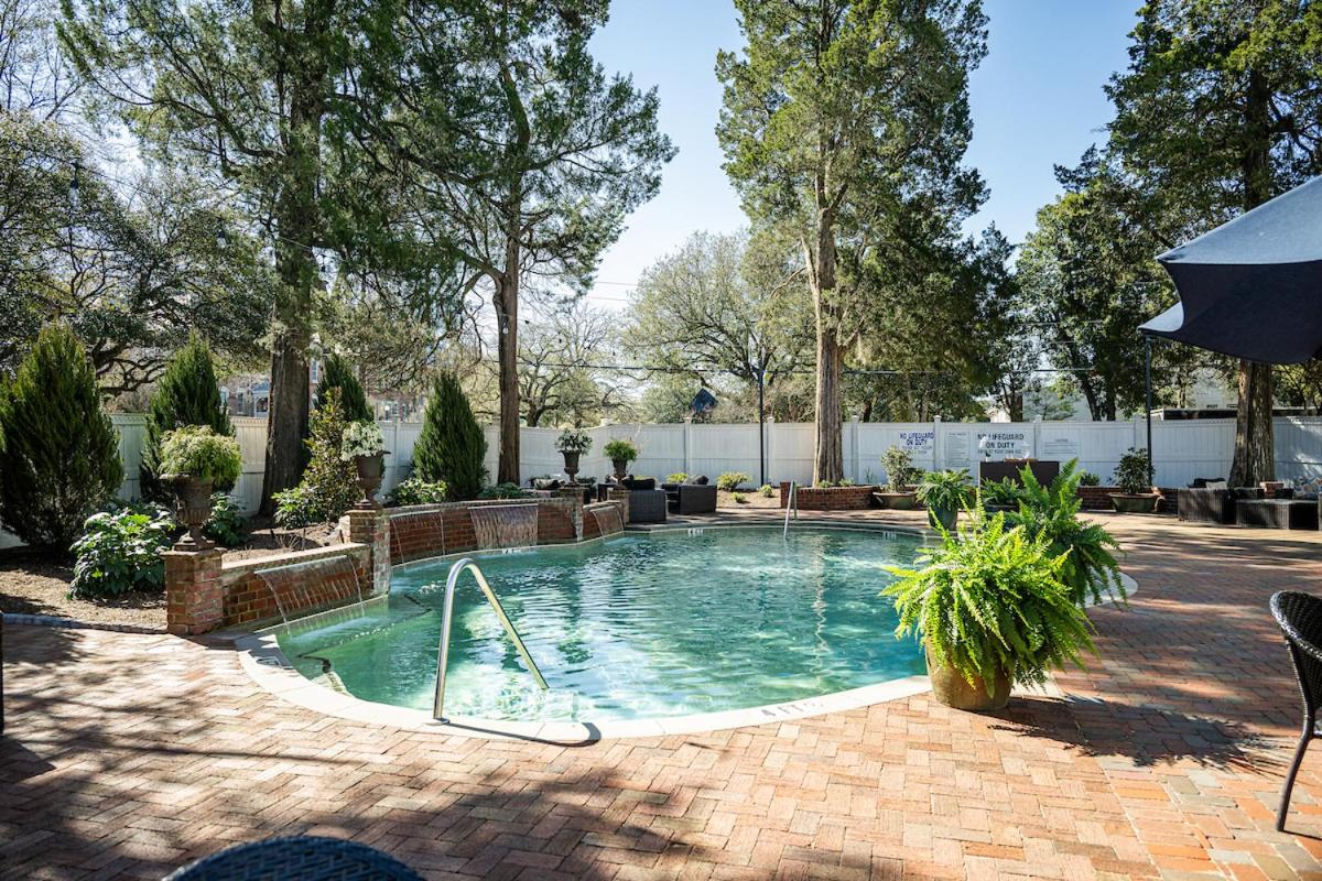 Outdoor swimming pool on a red brick patio, bordered by small brick waterfalls, potted ferns, and evergreen trees.