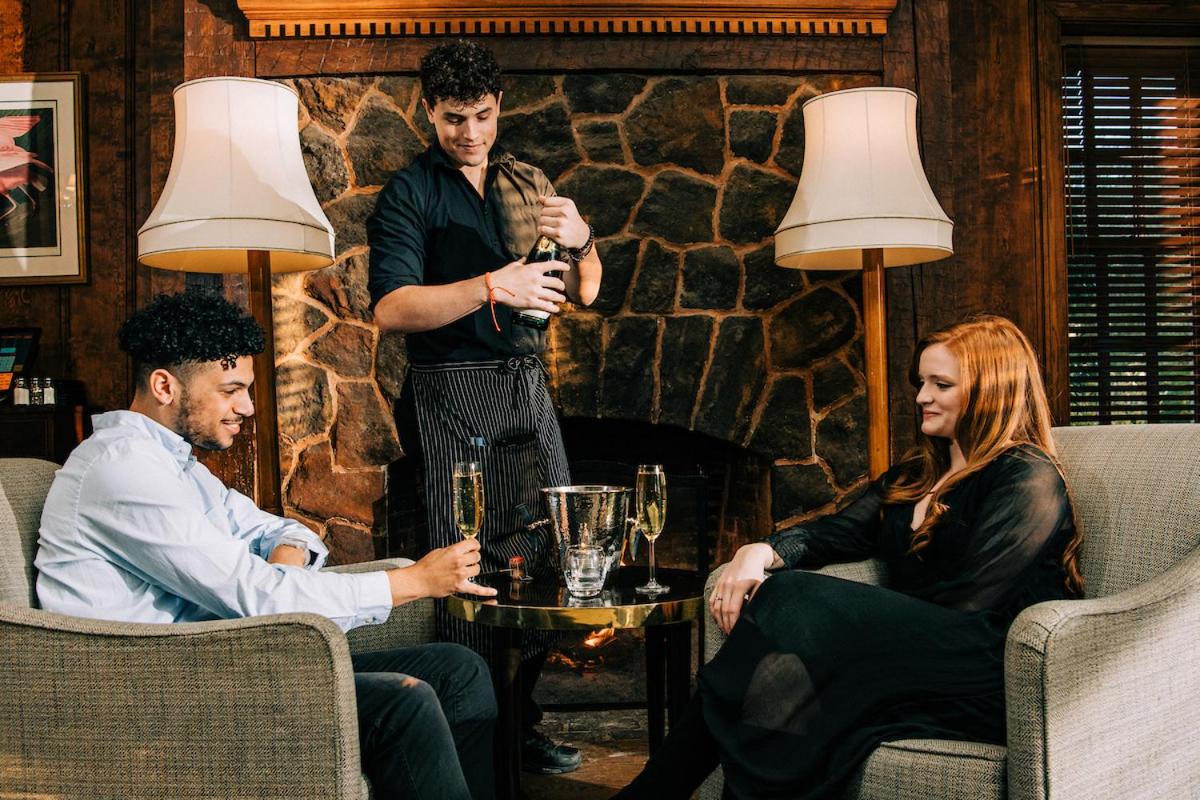 A server uncorks champagne behind patrons seated in armchairs before a rustic stone fireplace.