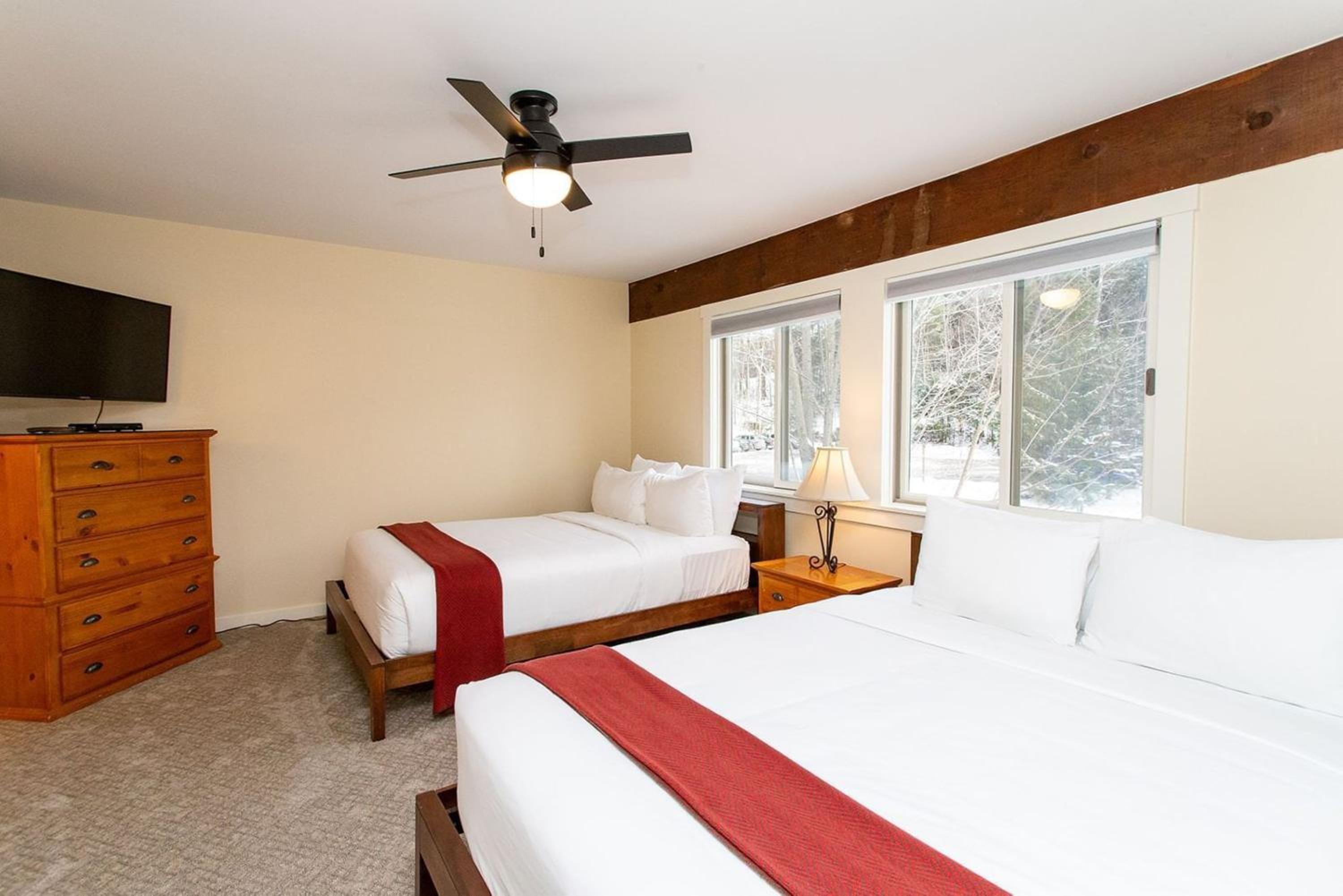 A bedroom with wood-frame beds, red throw blankets, a ceiling fan, dresser, and snowy woods through windows.