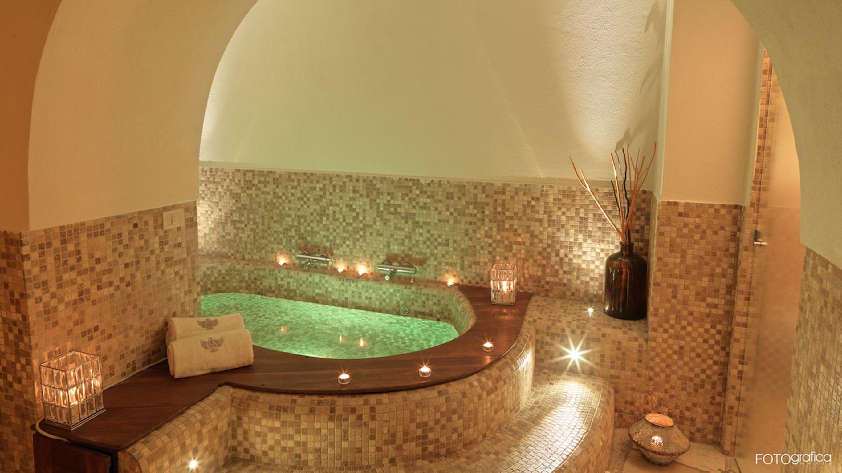 Sunken mosaic-tiled bath beneath a curved archway, surrounded by lit candles and warm lighting.