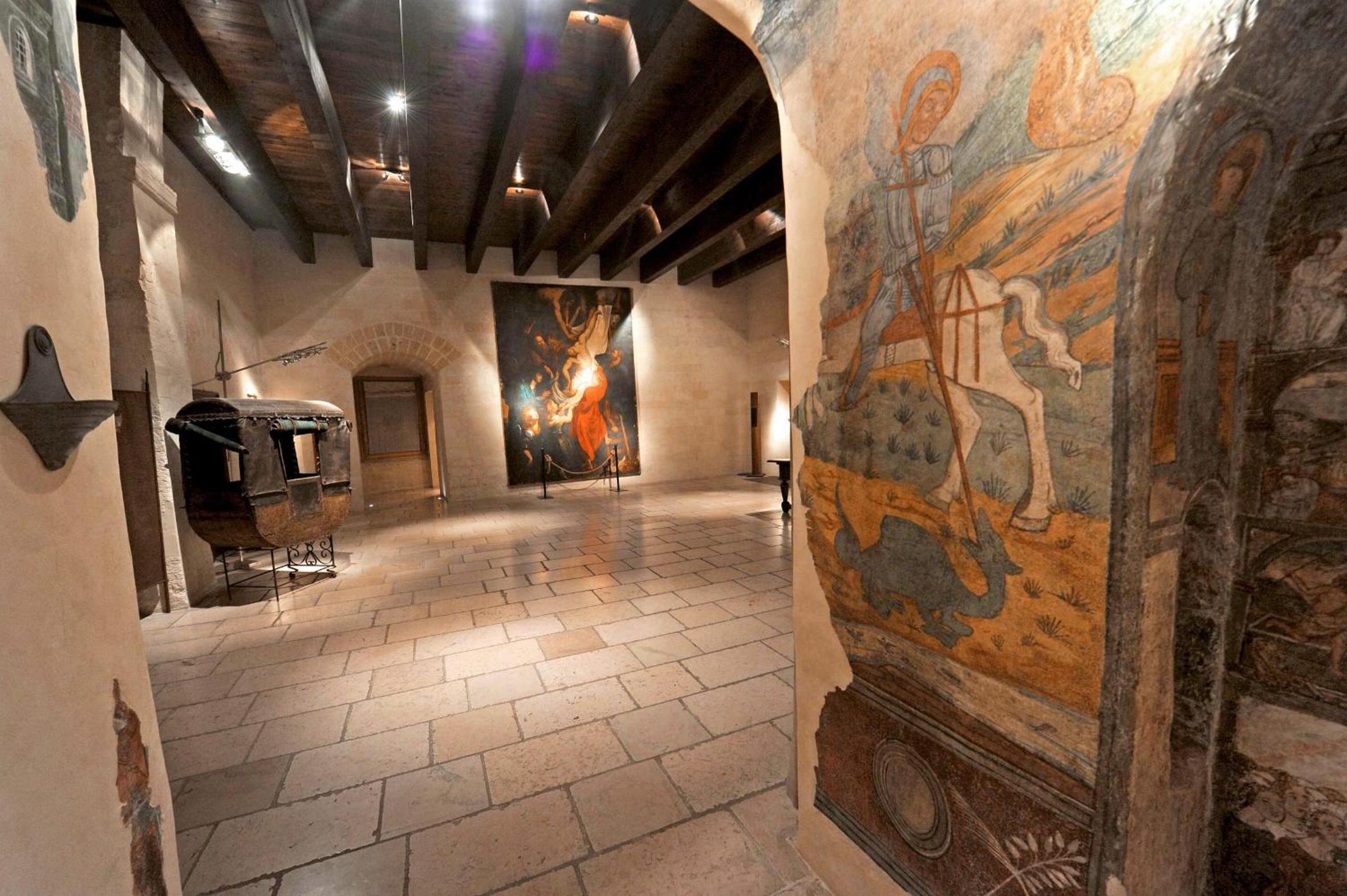 Stone hall with dark wooden ceiling beams, flagstone flooring, historic paintings, and a wall fresco.