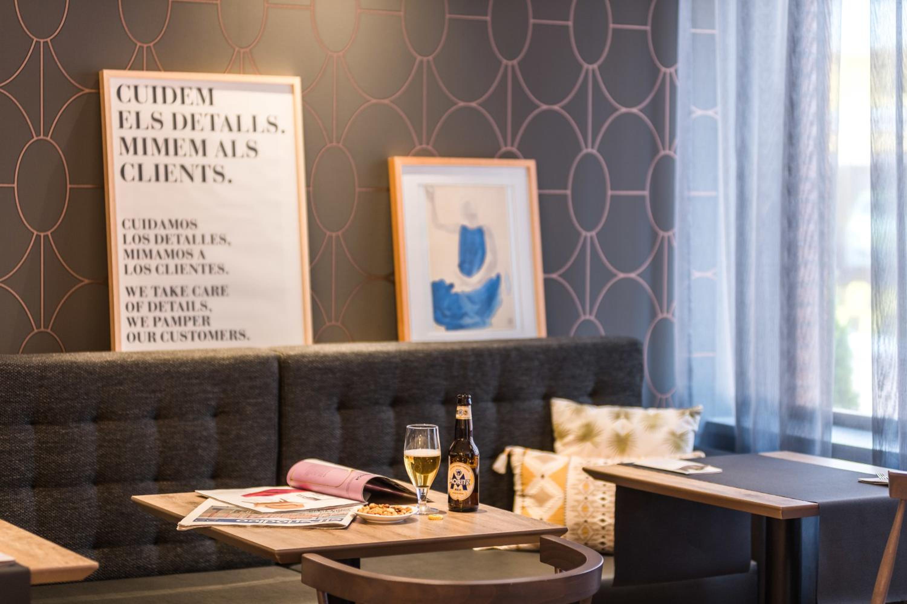 A dining nook with patterned wallpaper, framed art, a cushioned bench, and a drink on a wooden table.