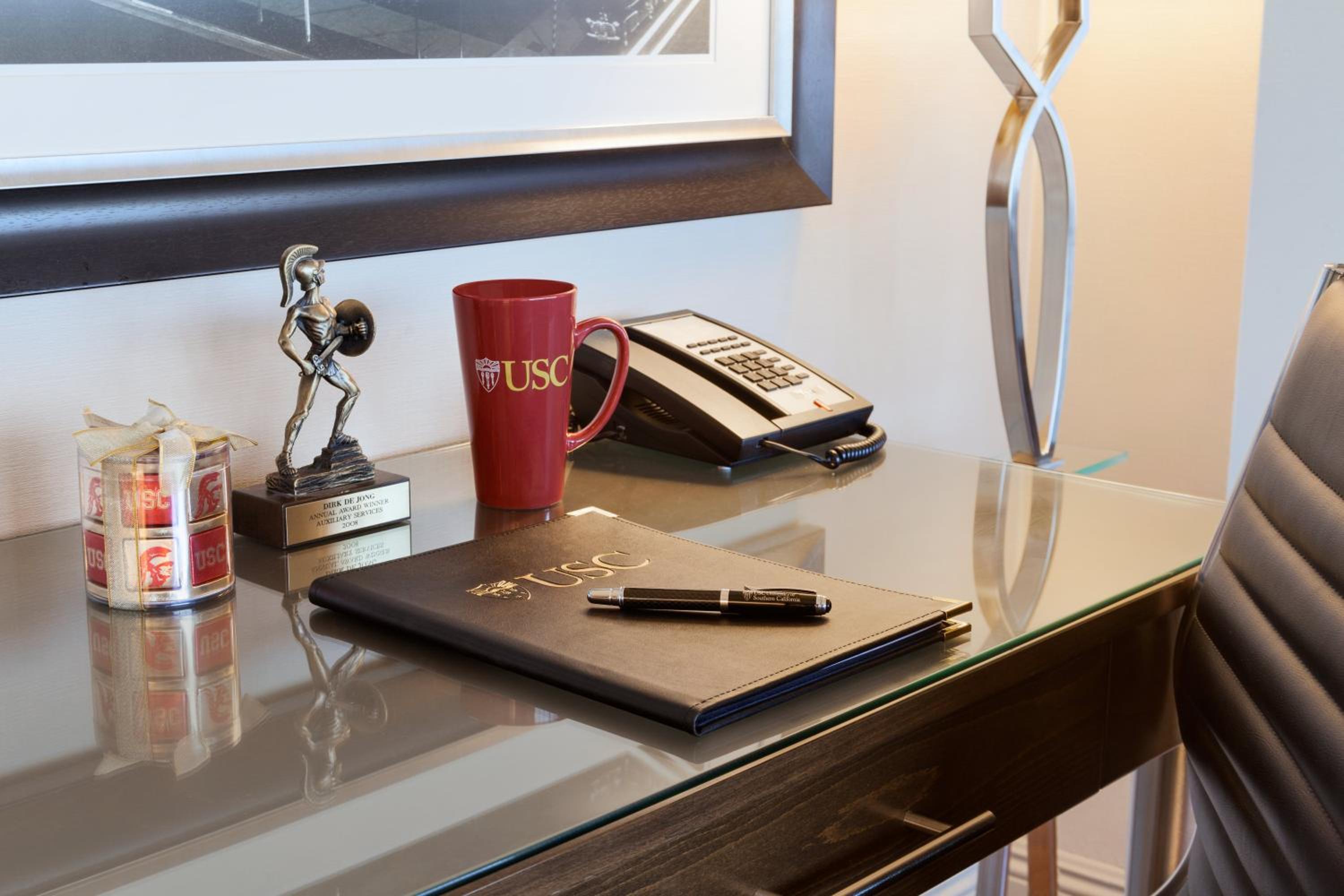 A hotel room desk with a leather-bound folder, USC mug, bronze mascot statuette, and telephone.