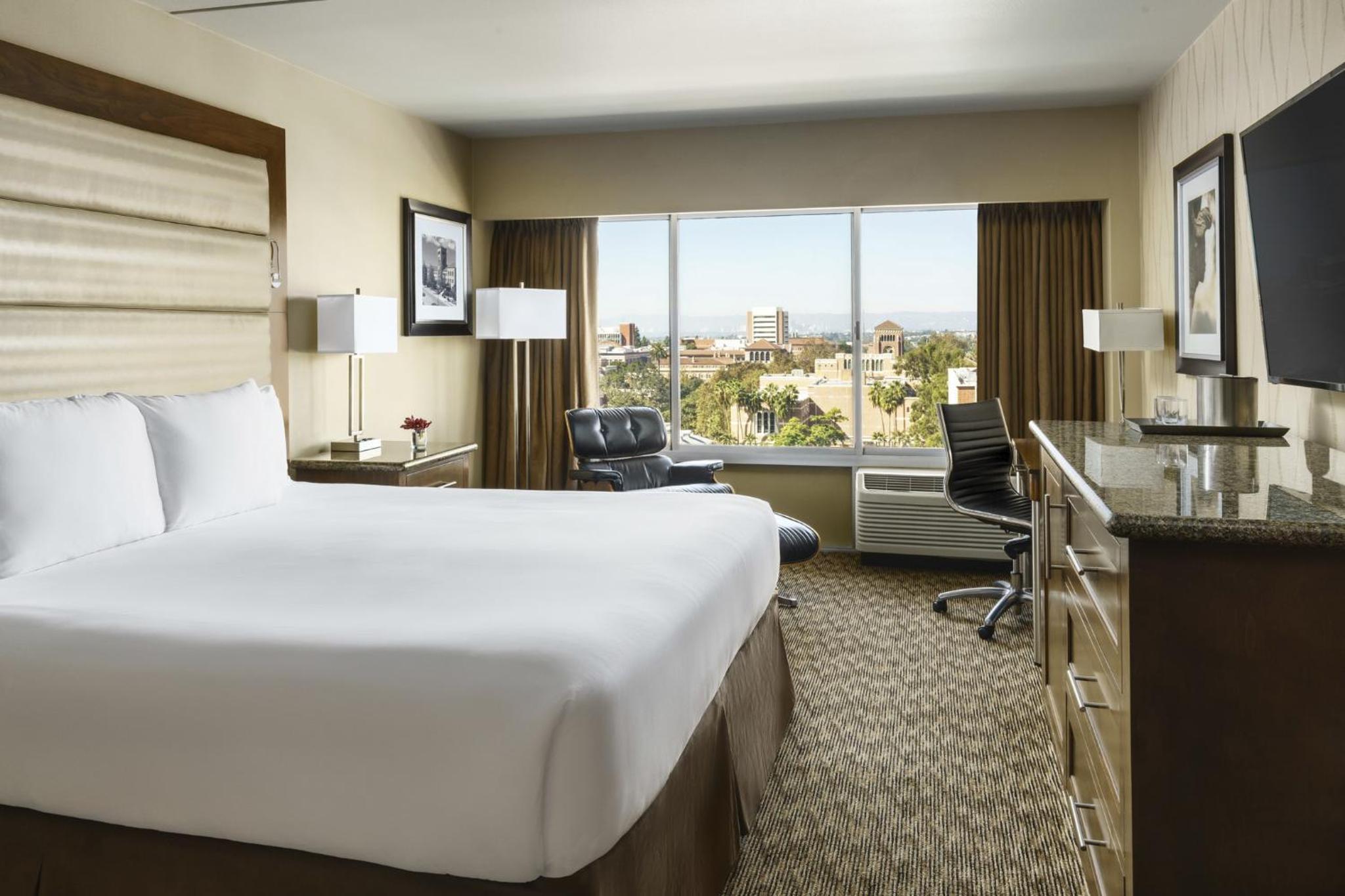 A guest bedroom with a king bed, leather lounge chair, and wide window overlooking the campus.