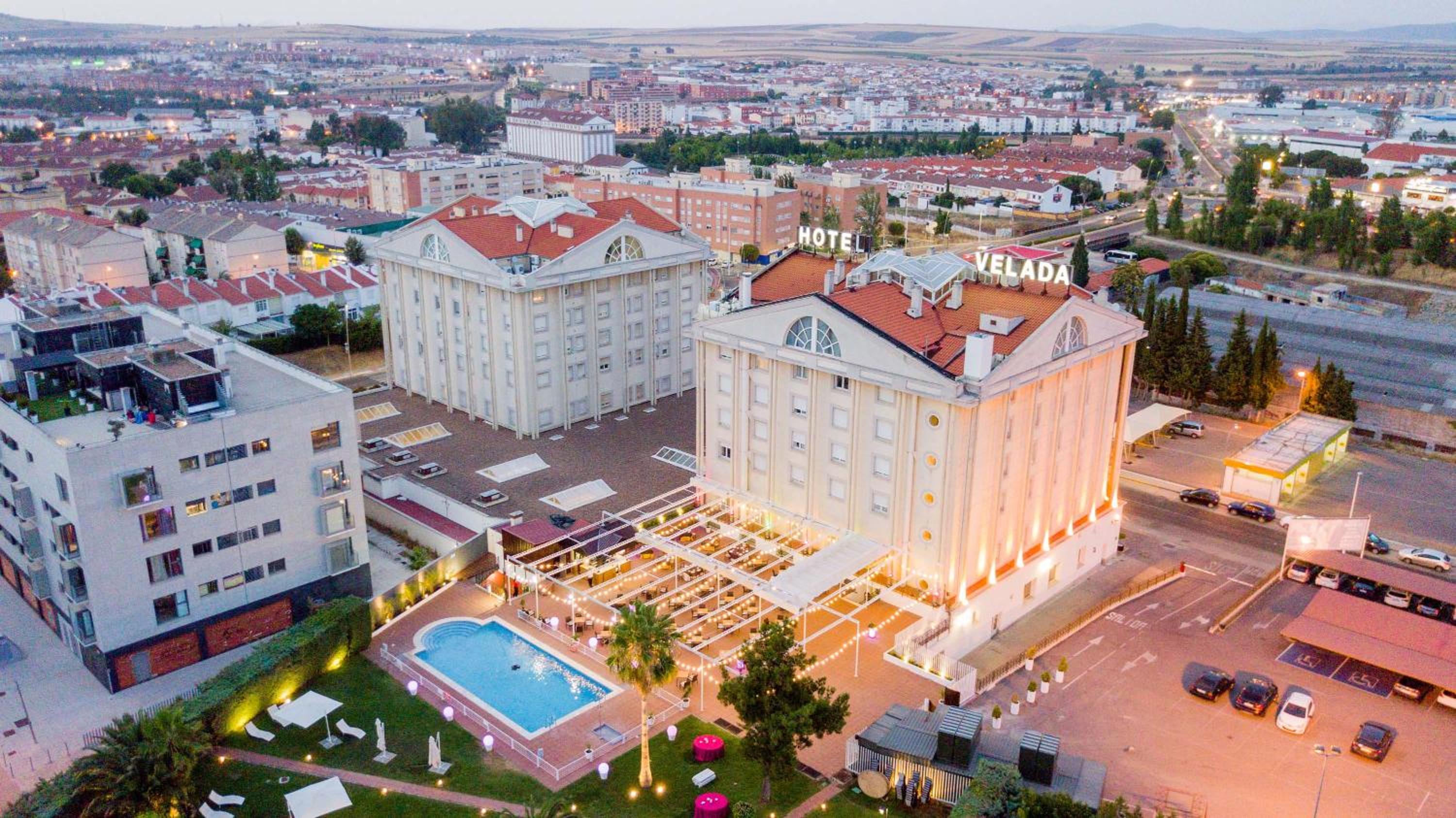 Aerial view at dusk of Velada Mérida hotel buildings with an illuminated terrace, swimming pool, and city beyond.