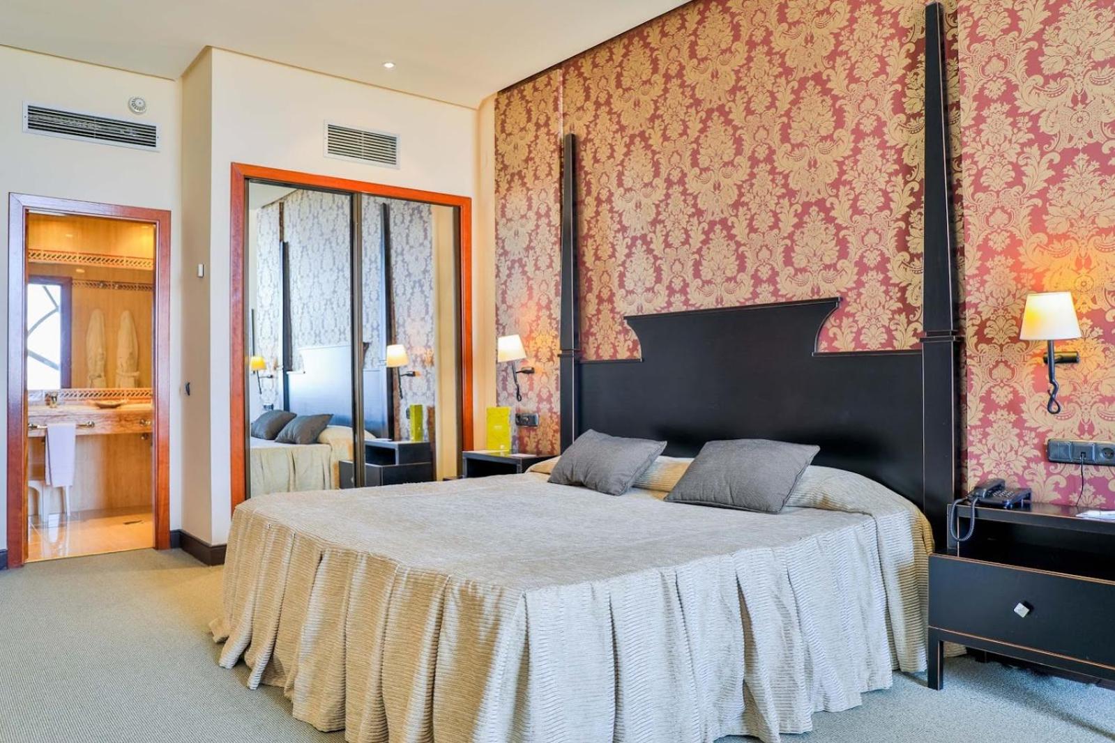 A hotel bedroom with patterned red wallpaper behind a tall dark headboard, a large bed, and an open bathroom door.