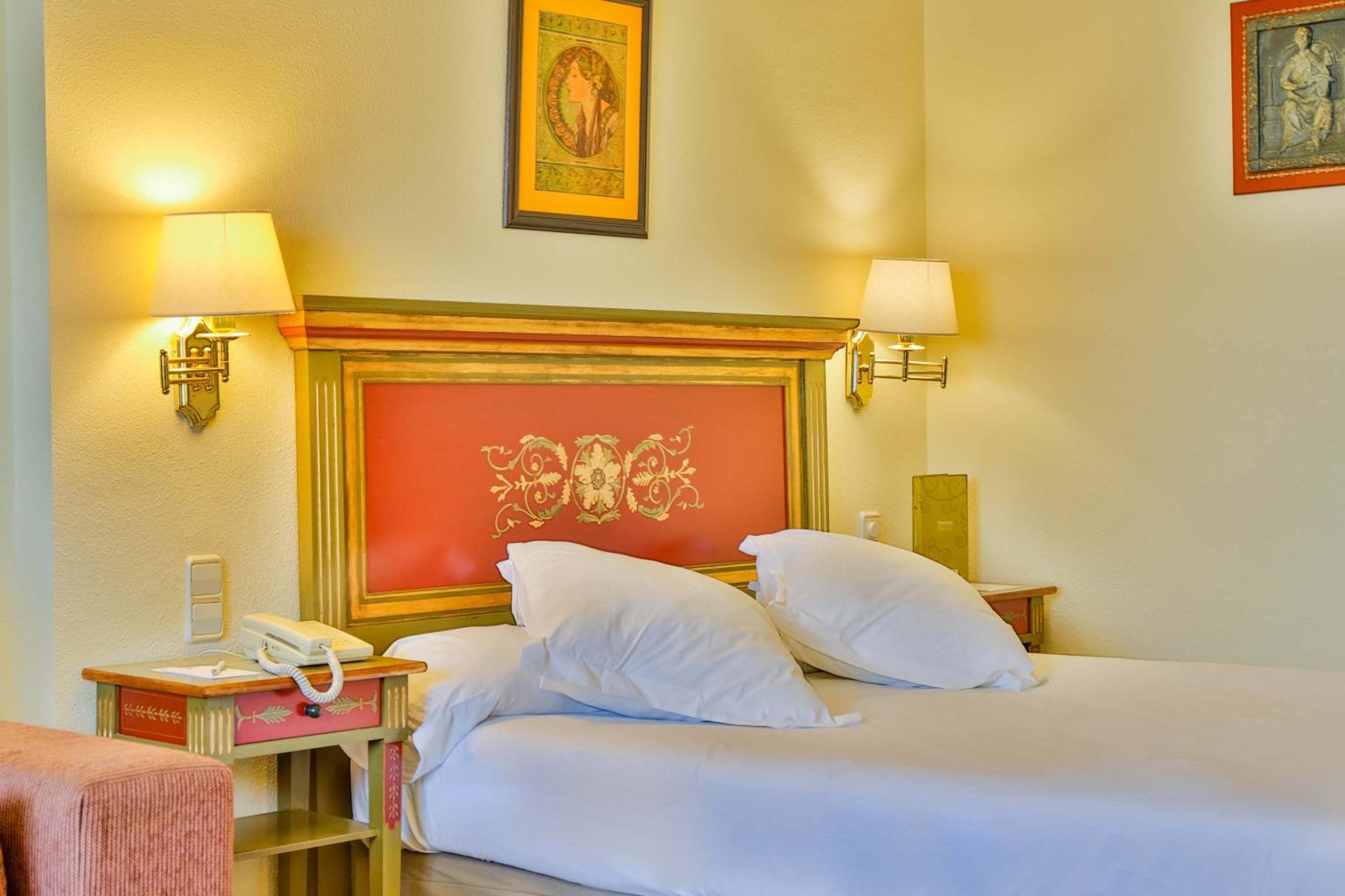 A hotel bed with white linens beneath a painted red and gold headboard, flanked by illuminated wall sconces.