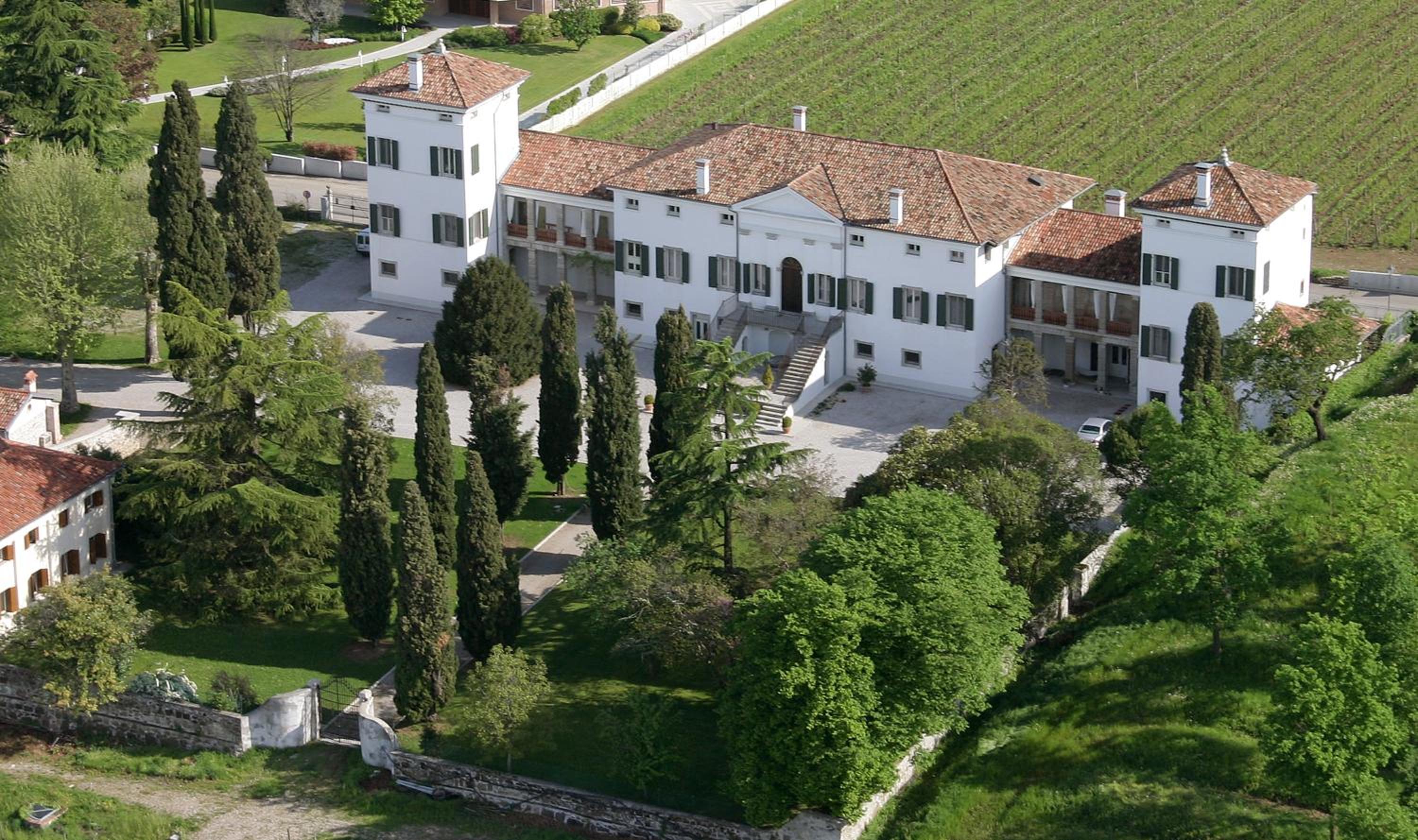 An aerial view of Villa Dragoni, showing a white villa with terracotta roofs, tall cypress trees, and green lawns.