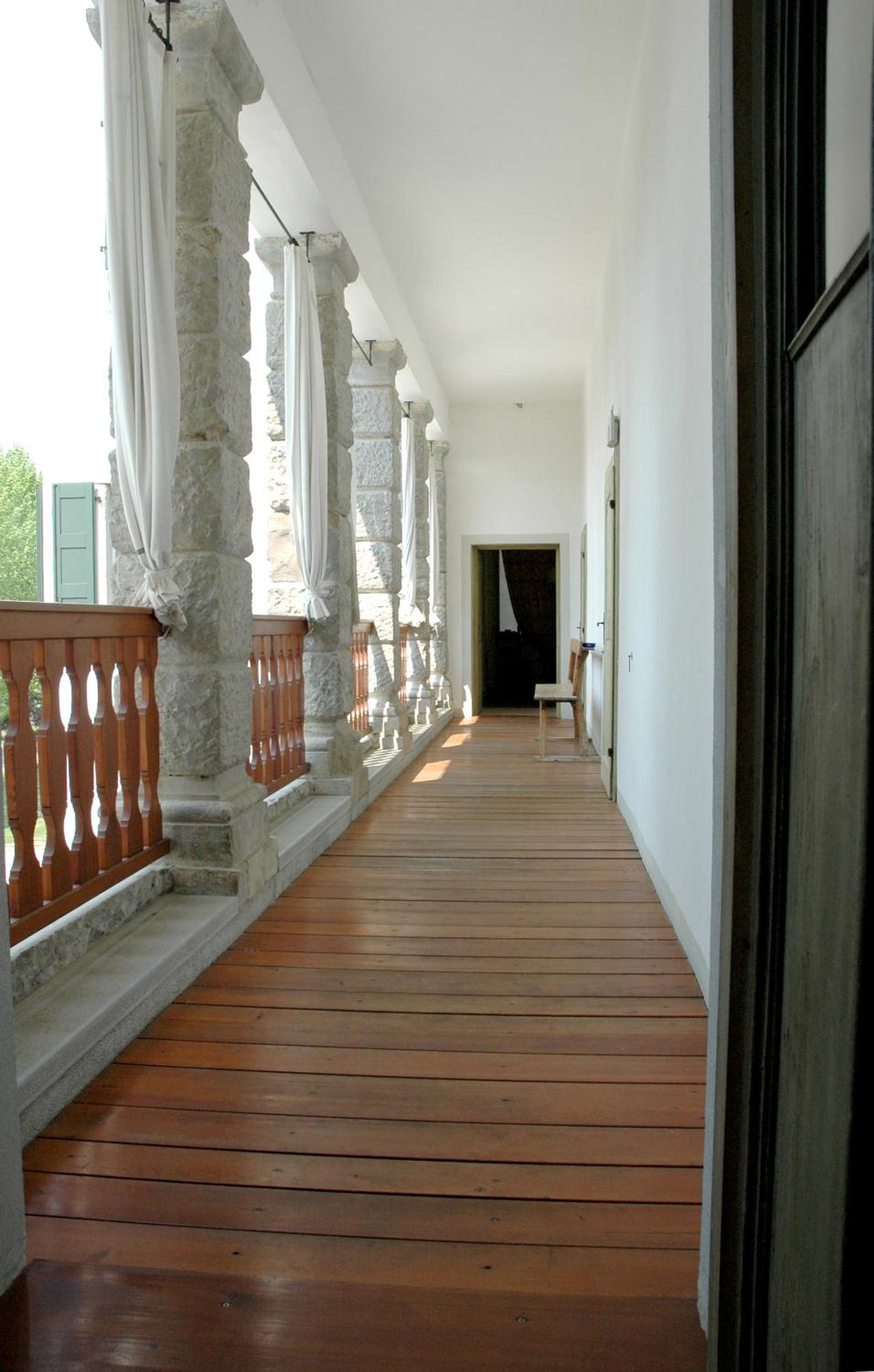 A covered loggia featuring rough stone pillars, a wooden balustrade, white curtains, and timber flooring.