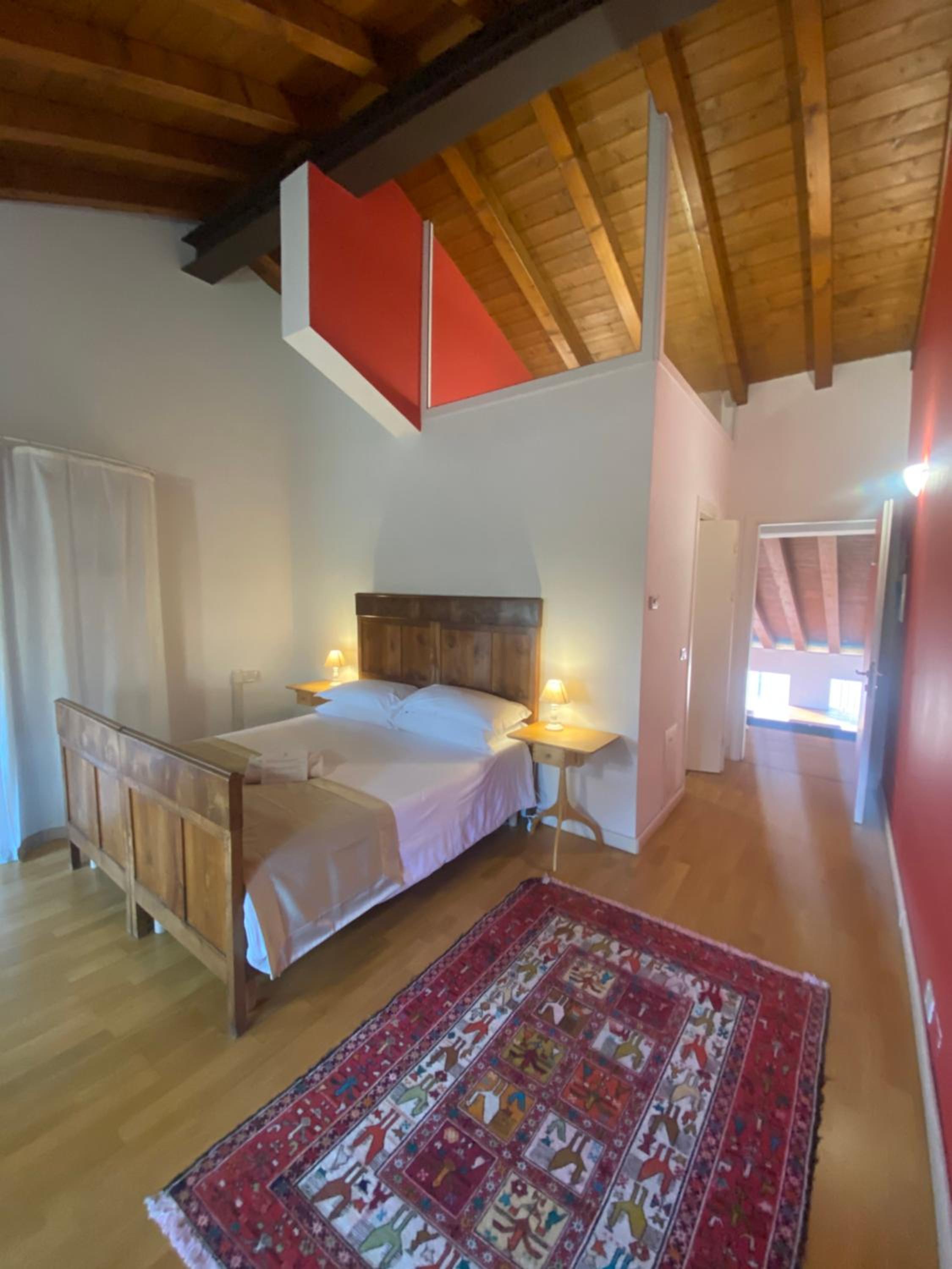 A bedroom with an exposed timber ceiling, wooden bed, red accent wall, and patterned rug on parquet flooring.