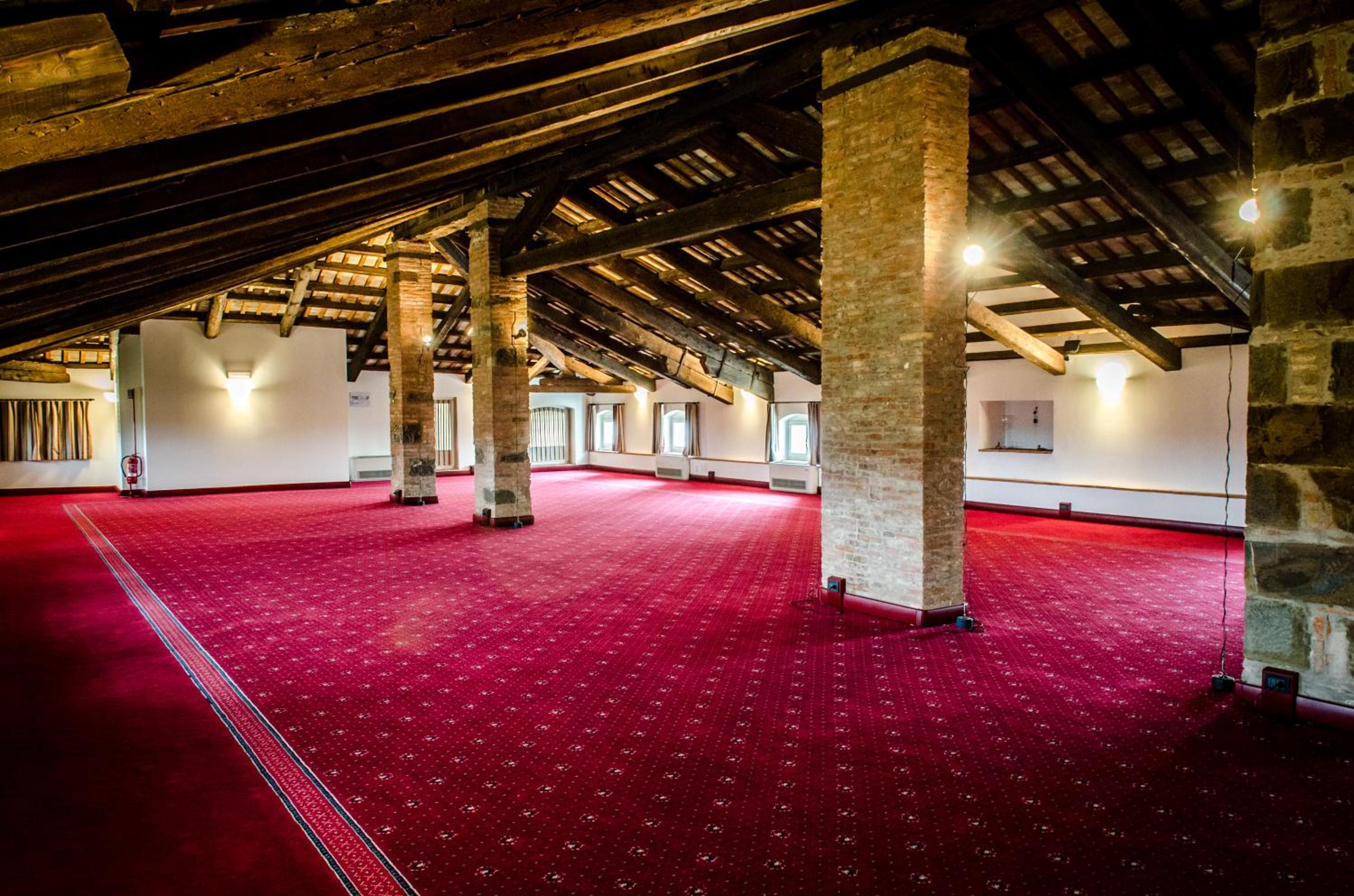 A spacious loft hall with exposed brick pillars, an open-beam wooden ceiling, and wall-to-wall red carpet.