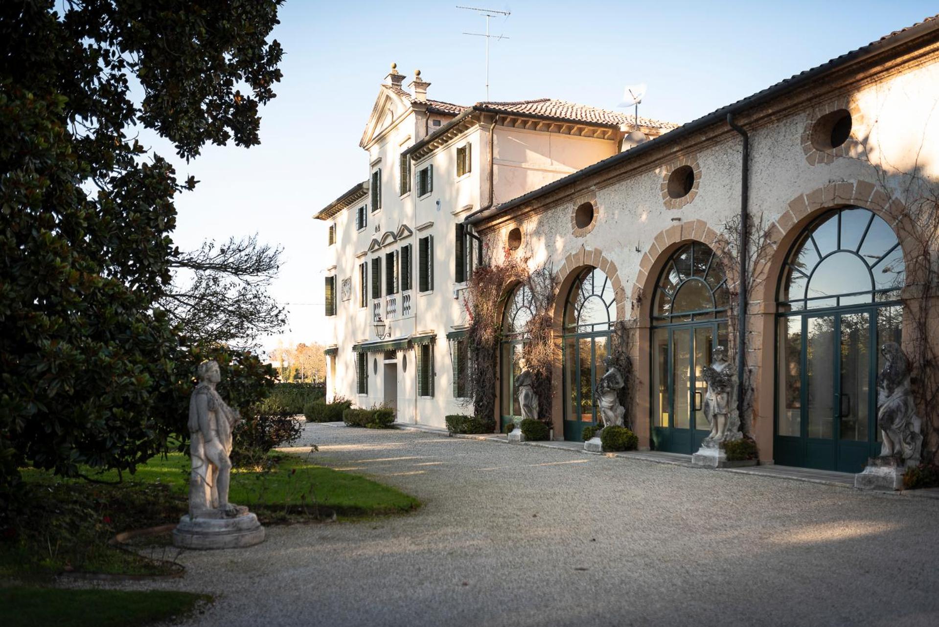 The exterior of Villa Vitturi featuring stone arches, classical statues, and a gravel driveway in sunlight.