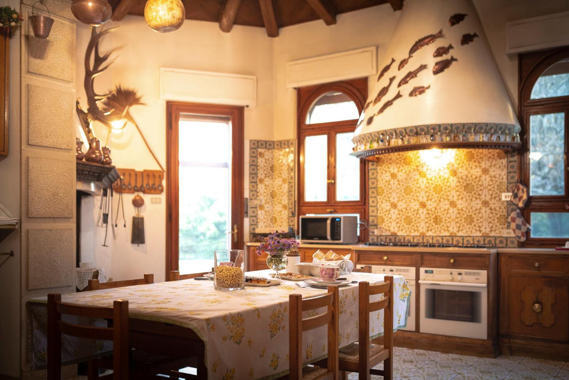 A rustic kitchen with patterned wall tiles, a conical range hood, and a dining table covered in floral cloth.