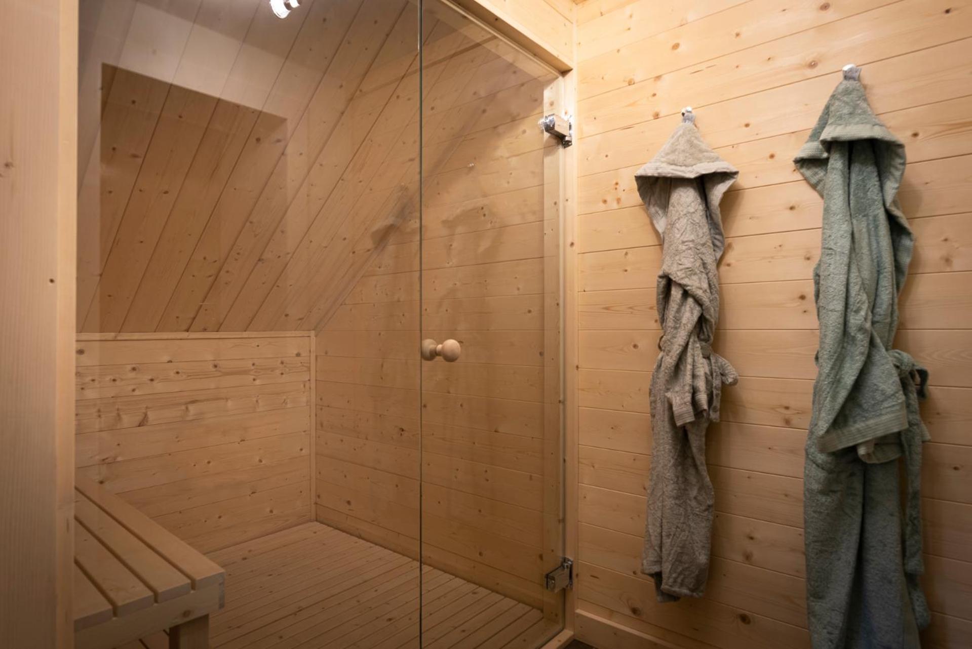 A wood-paneled sauna with a glass door and bathrobes hanging on wall hooks.