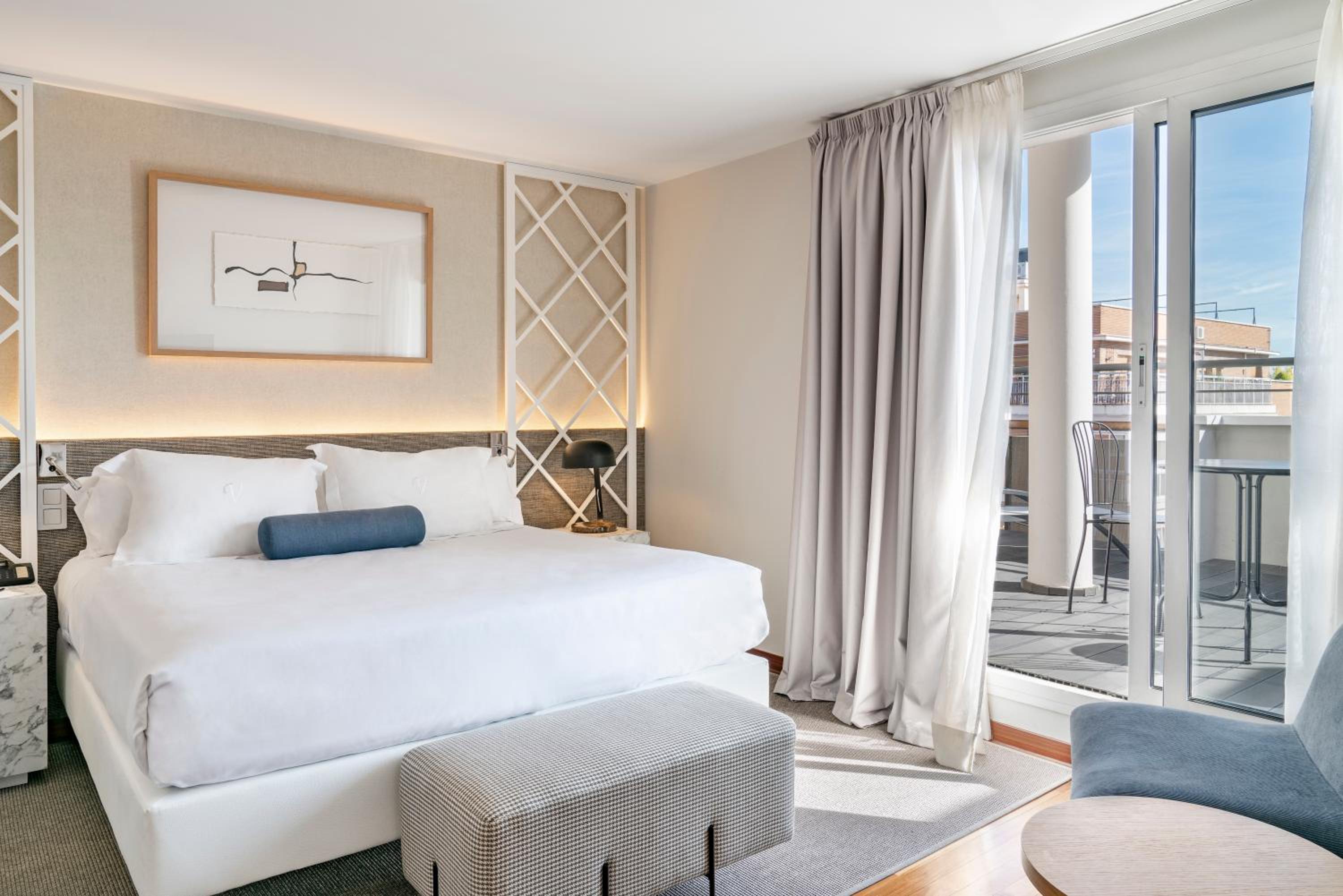 A hotel bedroom with a white made bed, an upholstered bench, and sliding doors opening to a sunlit terrace.