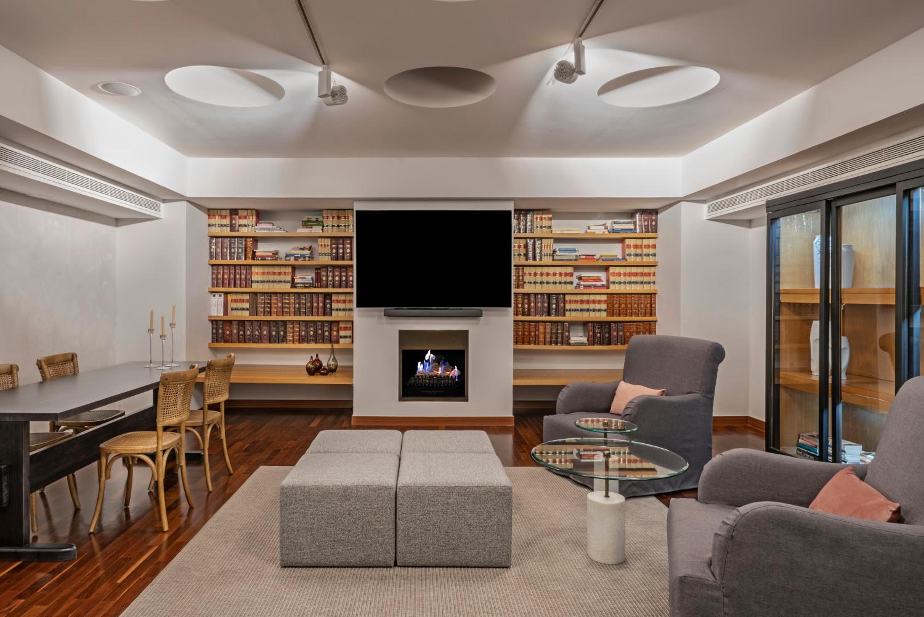 A lounge featuring hardwood floors, built-in bookshelves, armchairs, and a modern fireplace below a flat-screen TV.