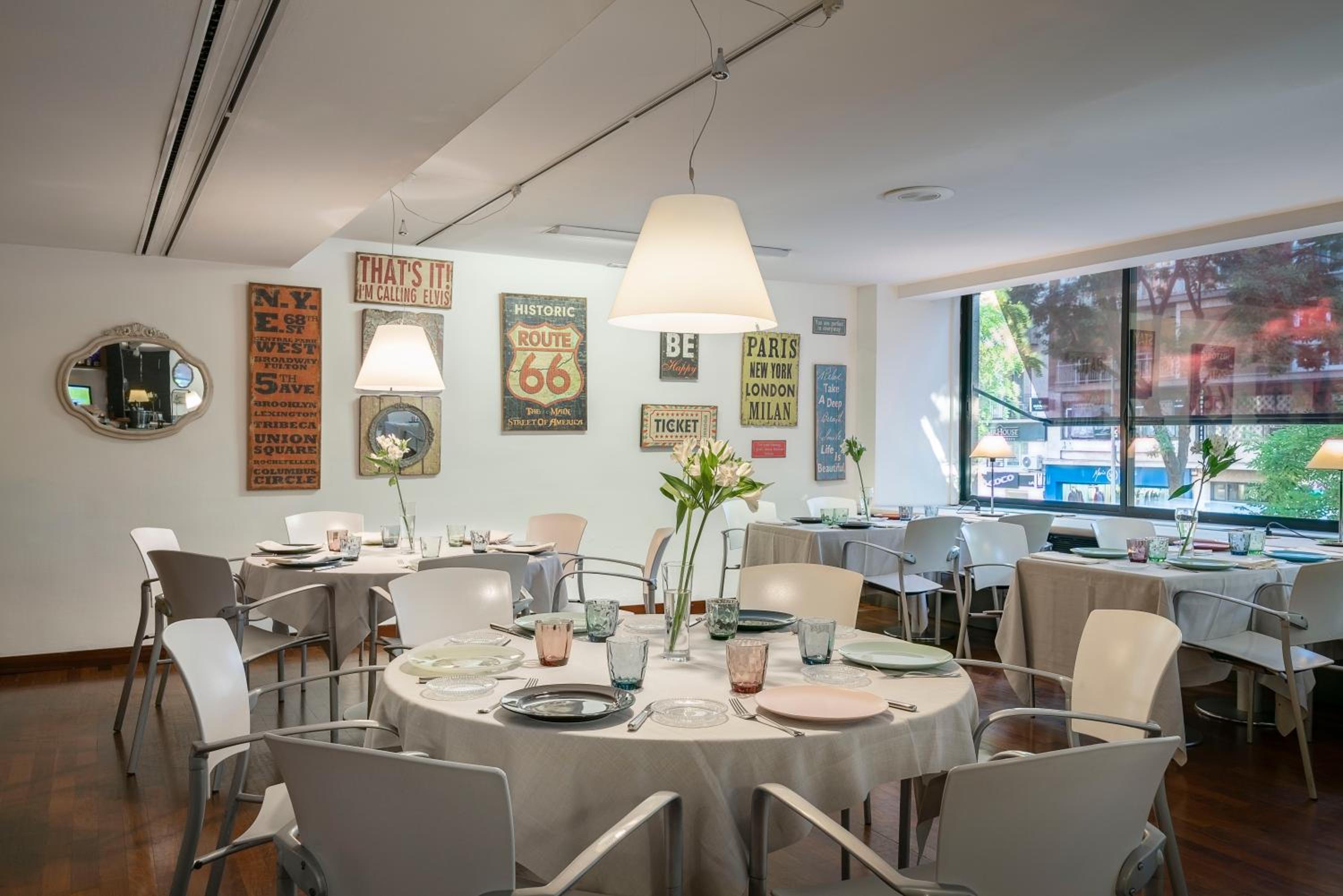 A bright dining room with round tables, neutral tablecloths, white chairs, and vintage posters along the white wall.