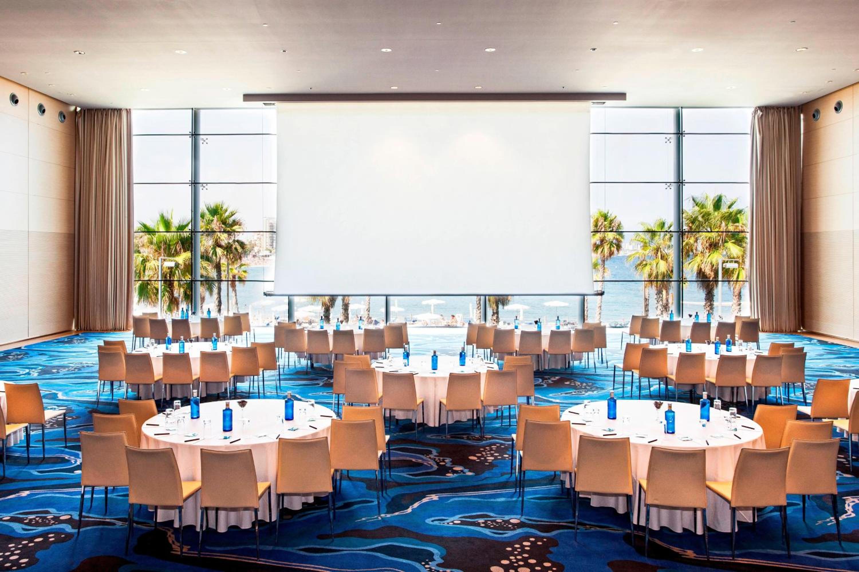 Banquet hall with round dining tables, blue patterned carpet, and large windows looking out to palm trees and the sea.