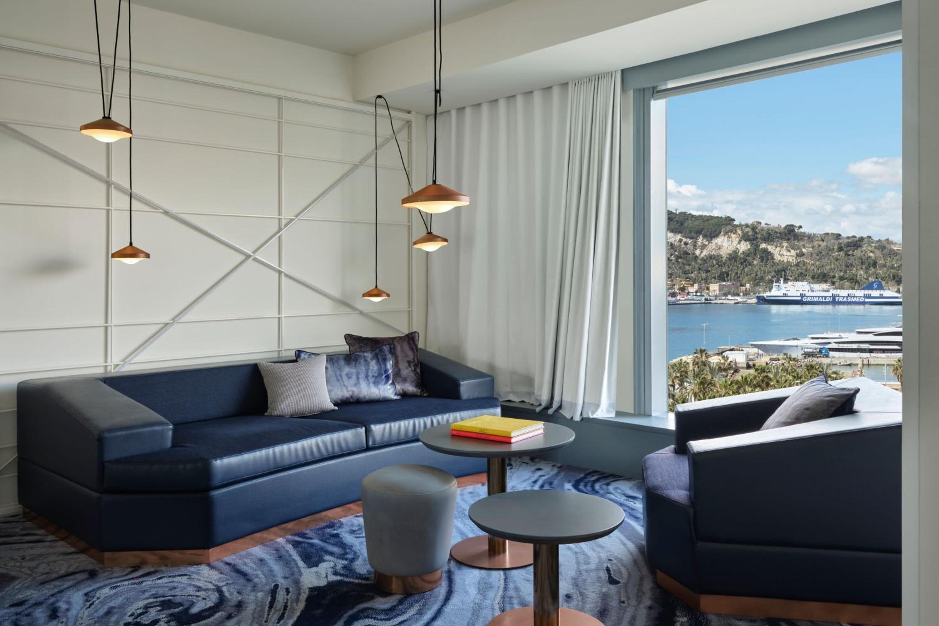 Hotel lounge area with a dark blue sofa, small tables, and a window overlooking the harbor and distant hills.