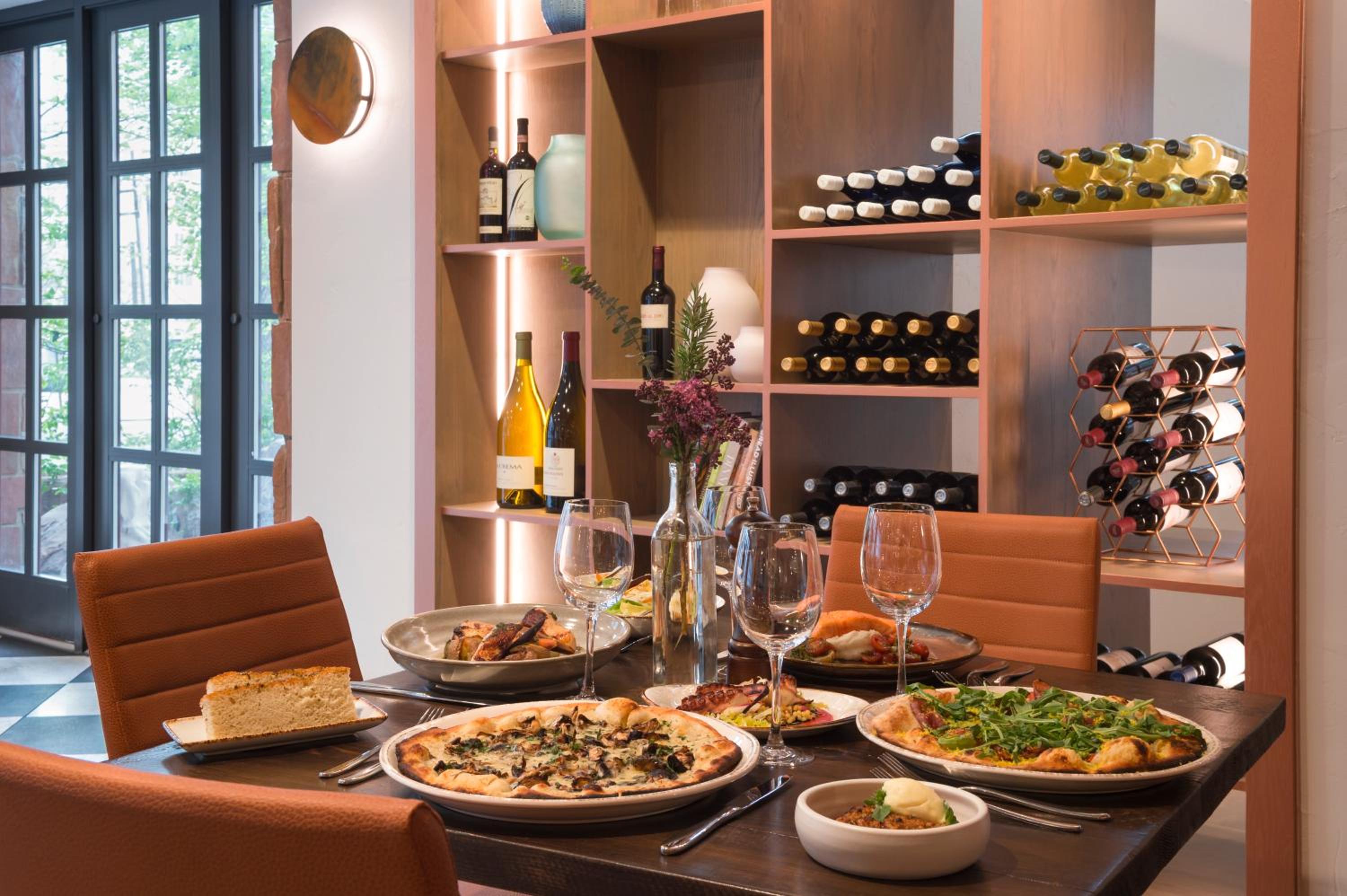 A dining table set with pizzas, wine glasses, and dishes in front of built-in wooden wine shelving.