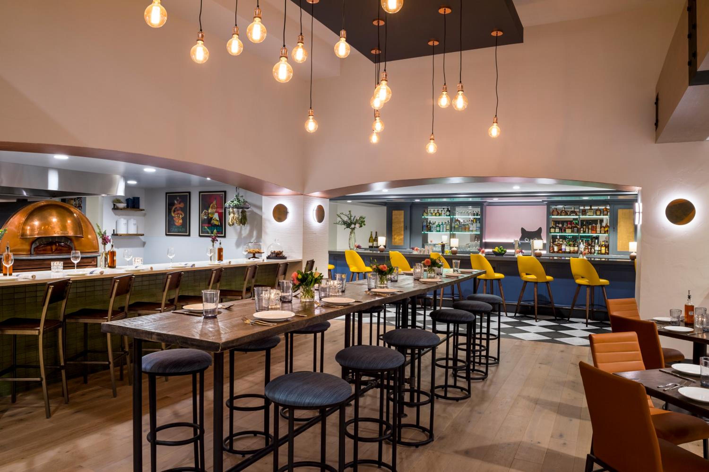 A restaurant dining area with long high-top tables, pendant bulbs, a copper oven, and a bar counter.