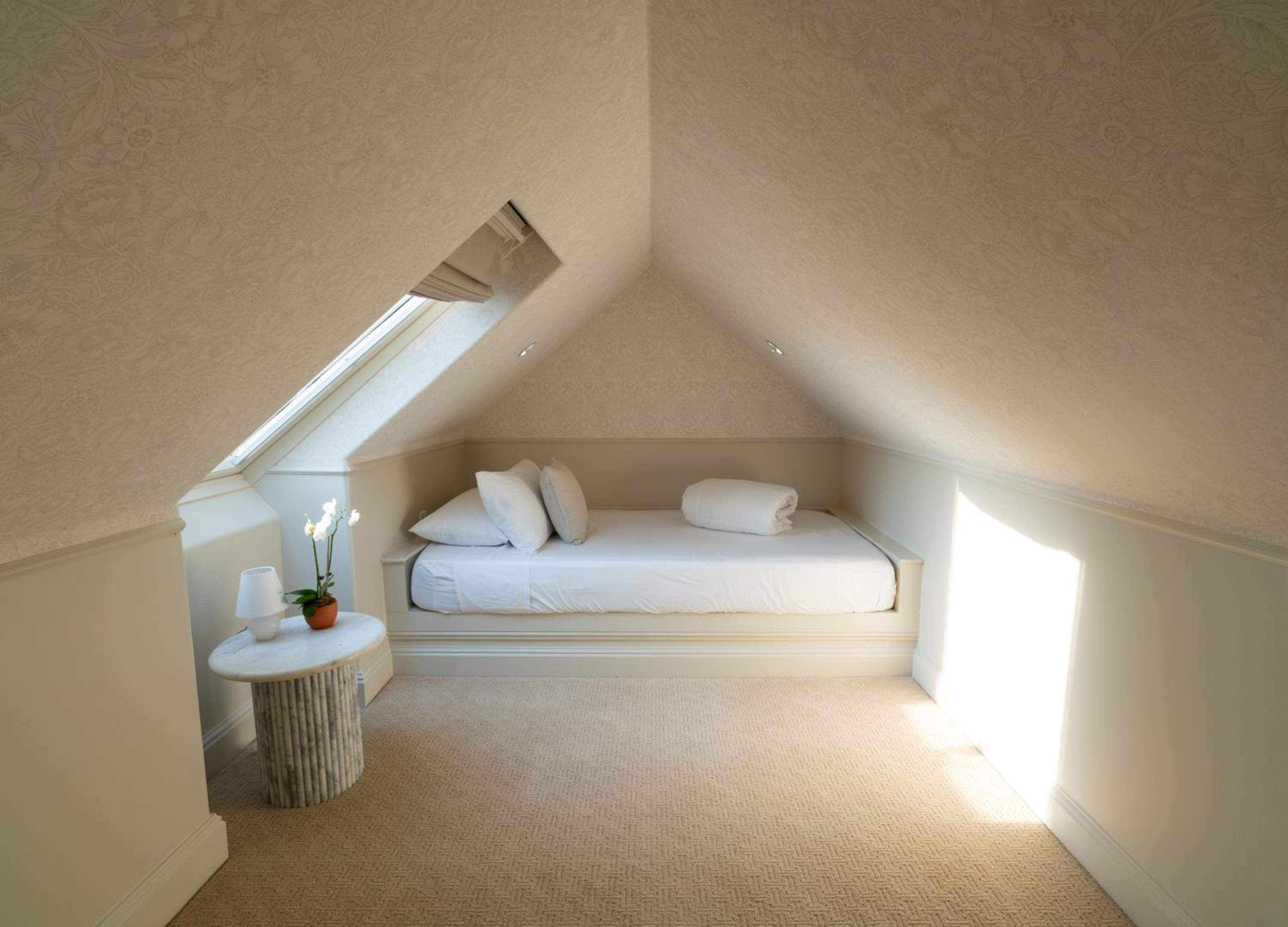 A bed is built into an alcove beneath the pitched ceiling and skylight of a wallpapered attic room.
