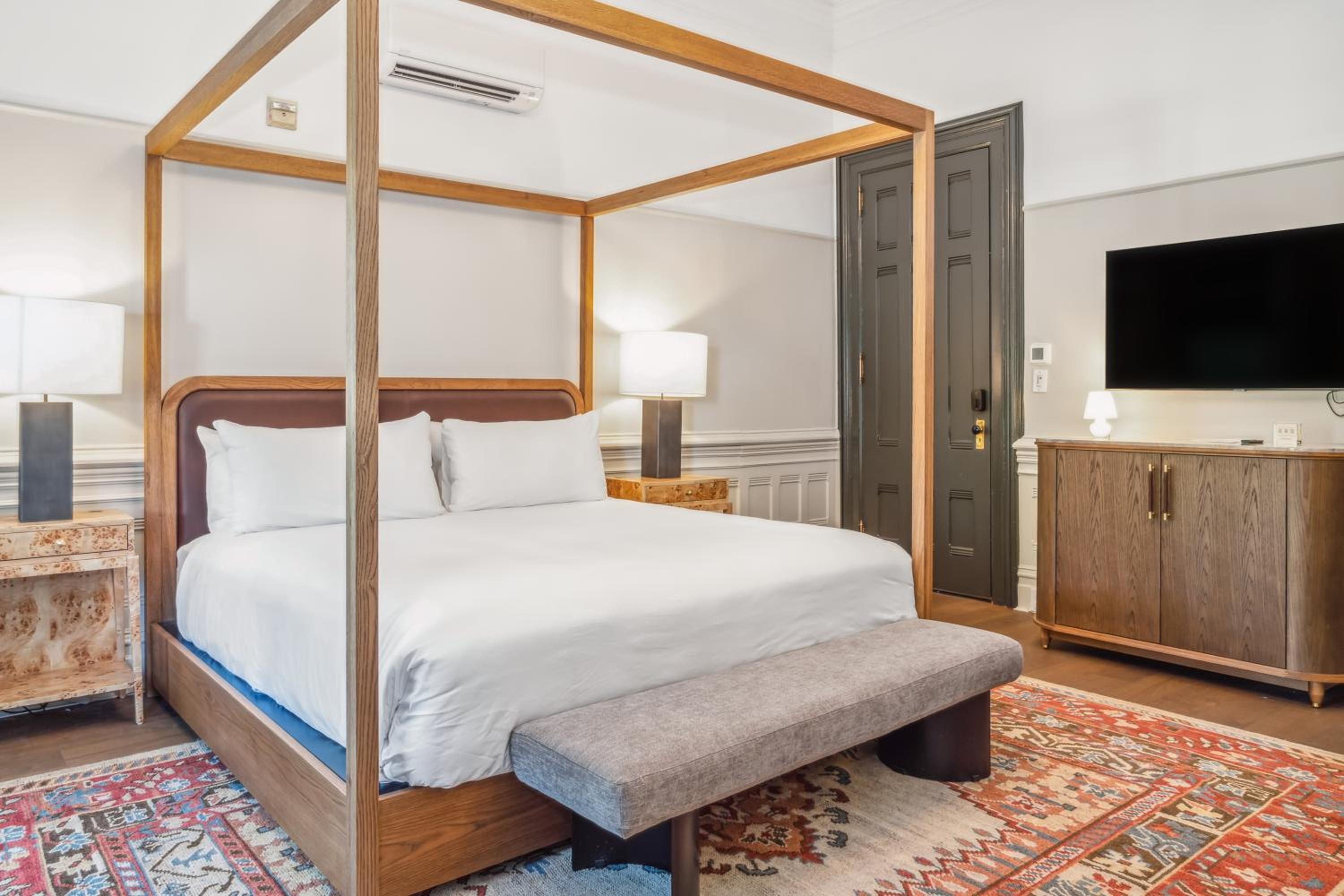 A wooden canopy bed rests on an intricate patterned rug in a bedroom with a dark wood door.