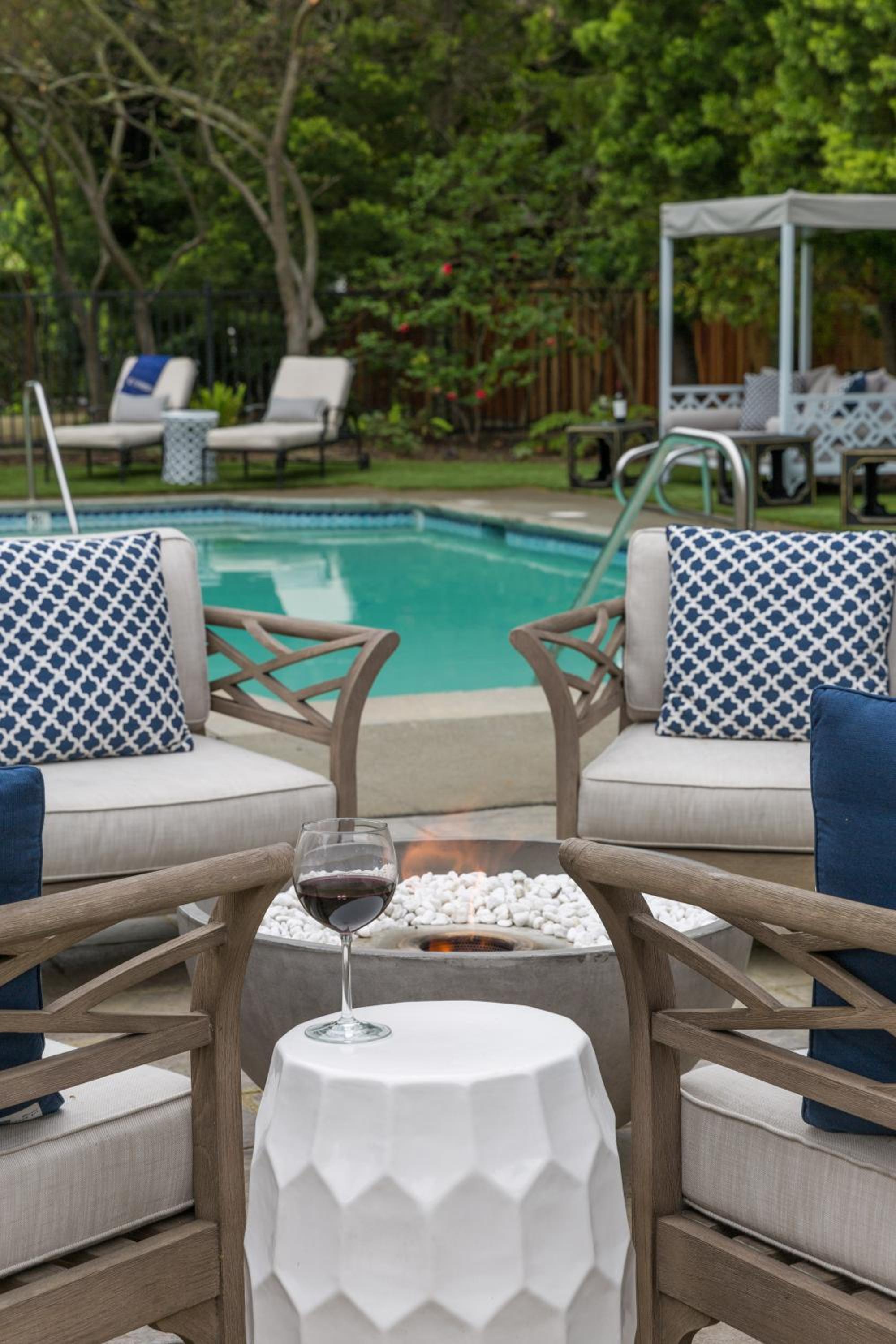 A glass of red wine sits on a fluted pedestal table beside an outdoor fire pit and swimming pool.