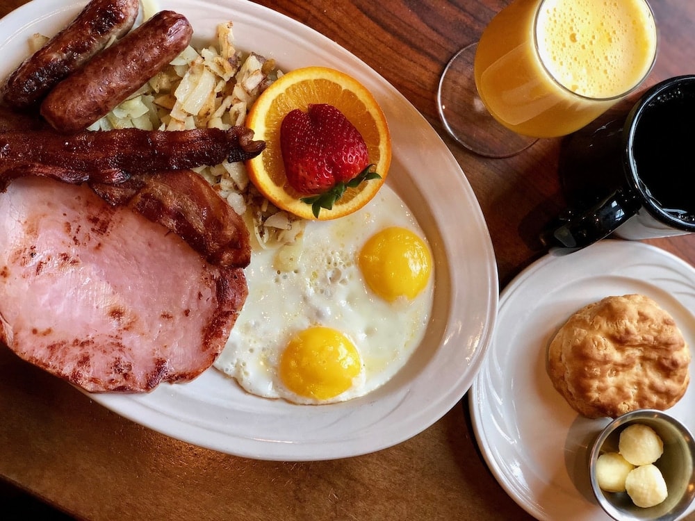A breakfast plate with fried eggs, ham, bacon, sausage, and hash browns served beside juice and a biscuit.