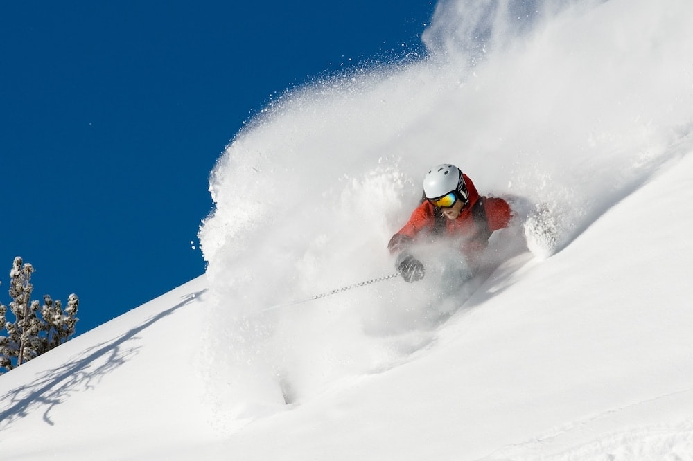 A skier in a red jacket maneuvers through deep powder snow on a sunlit slope beneath a blue sky.