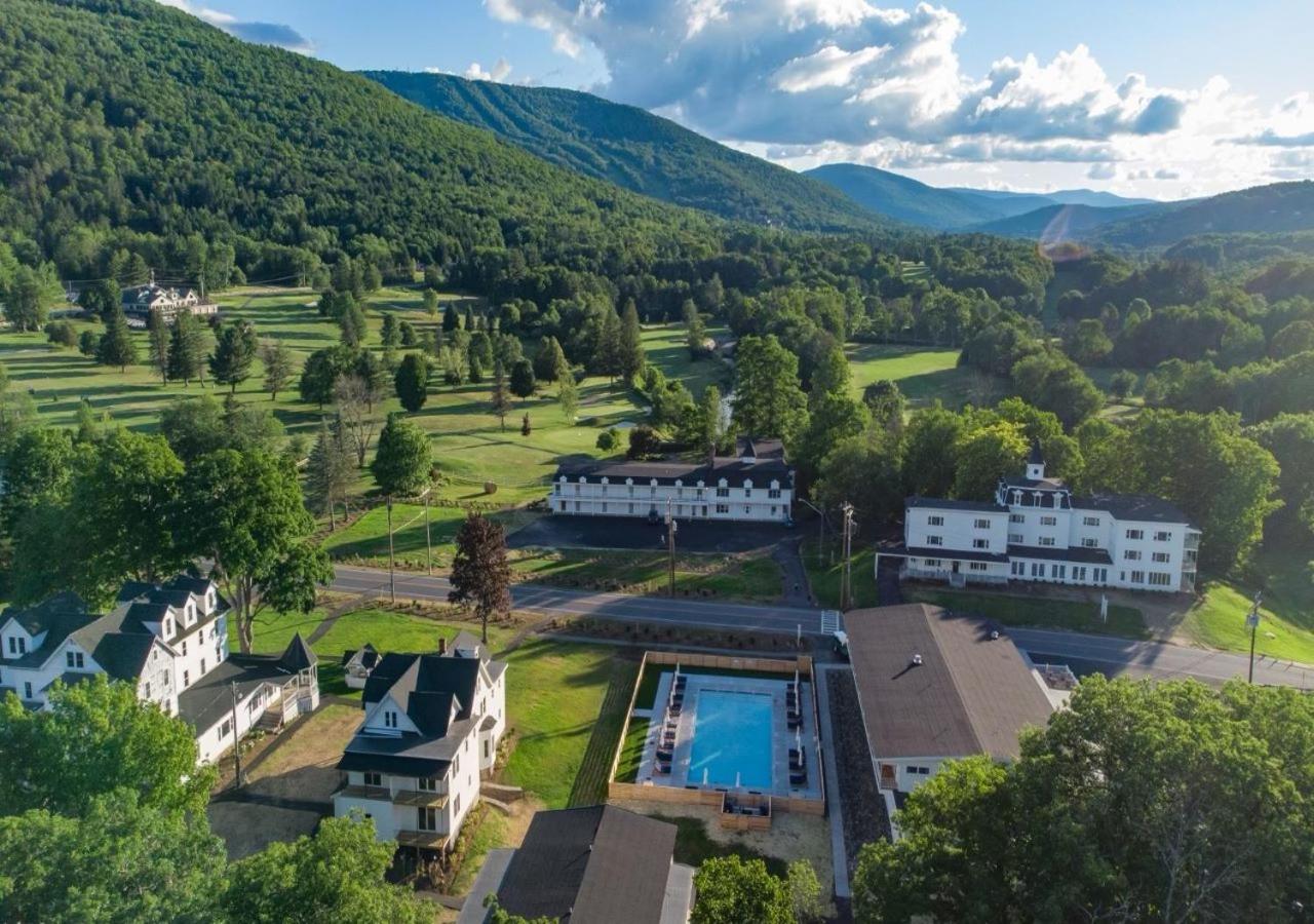 Aerial view of resort buildings, a swimming pool, and a green golf course set against wooded mountains.